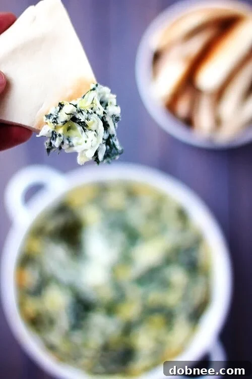 A close-up shot of creamy Spinach Artichoke Dip in a white bowl, with dippers around it.