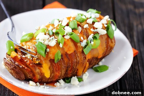 Buffalo Hasselback Potatoes, garnished with blue cheese and green onions, resting on a baking sheet, with hot sauce in the background.