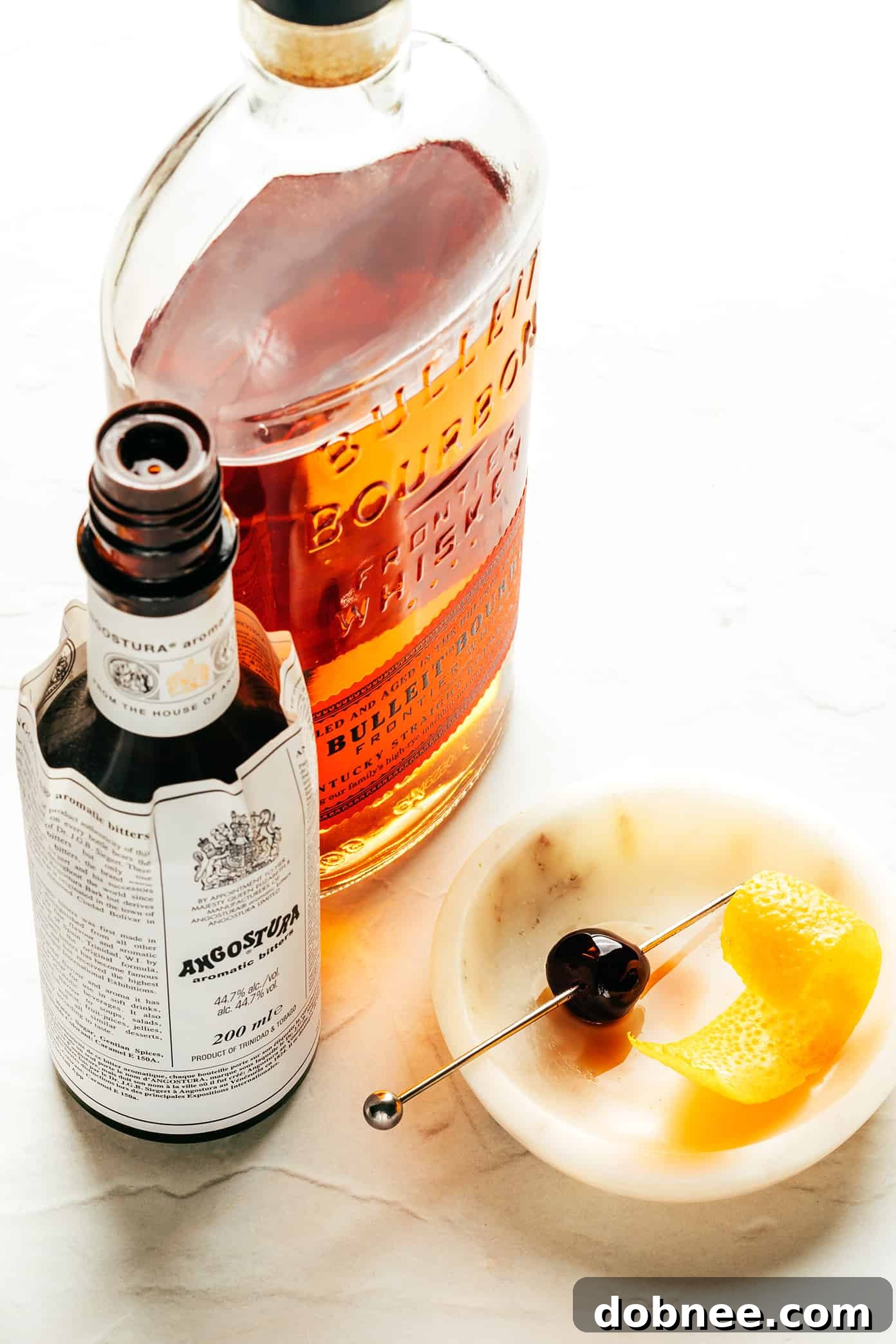 Fresh ingredients for an Old Fashioned cocktail including a bottle of bourbon, a jar of bitters, an orange, sugar cubes, and a jar of Luxardo cherries.