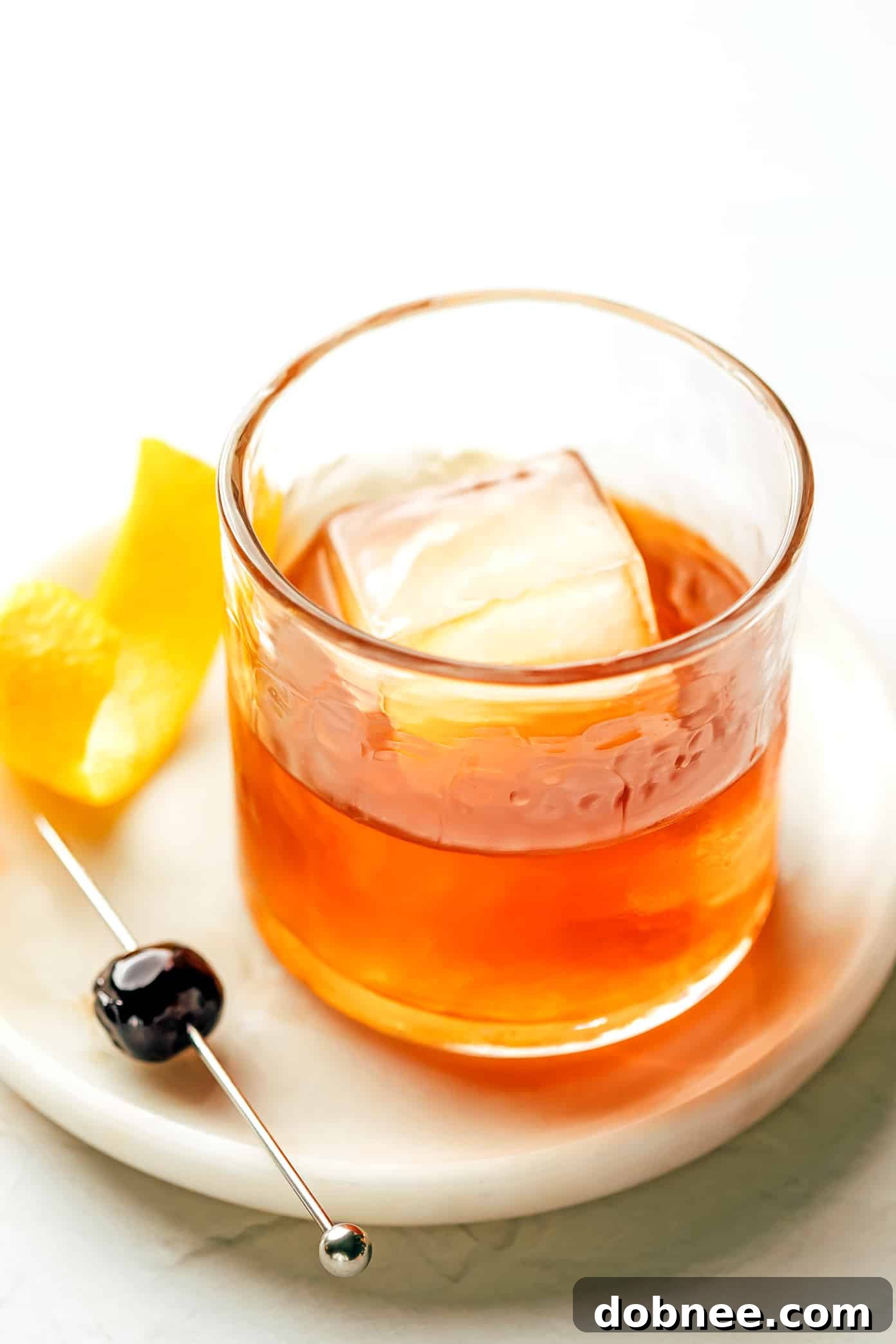 A close-up of a beautifully presented Old Fashioned cocktail with a large ice cube, a twisted orange peel, and a dark Luxardo cherry inside a crystal lowball glass.