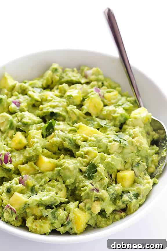 A close-up of a spoon mashing ripe avocados in a bowl, with pineapple chunks visible, for making pineapple guacamole.