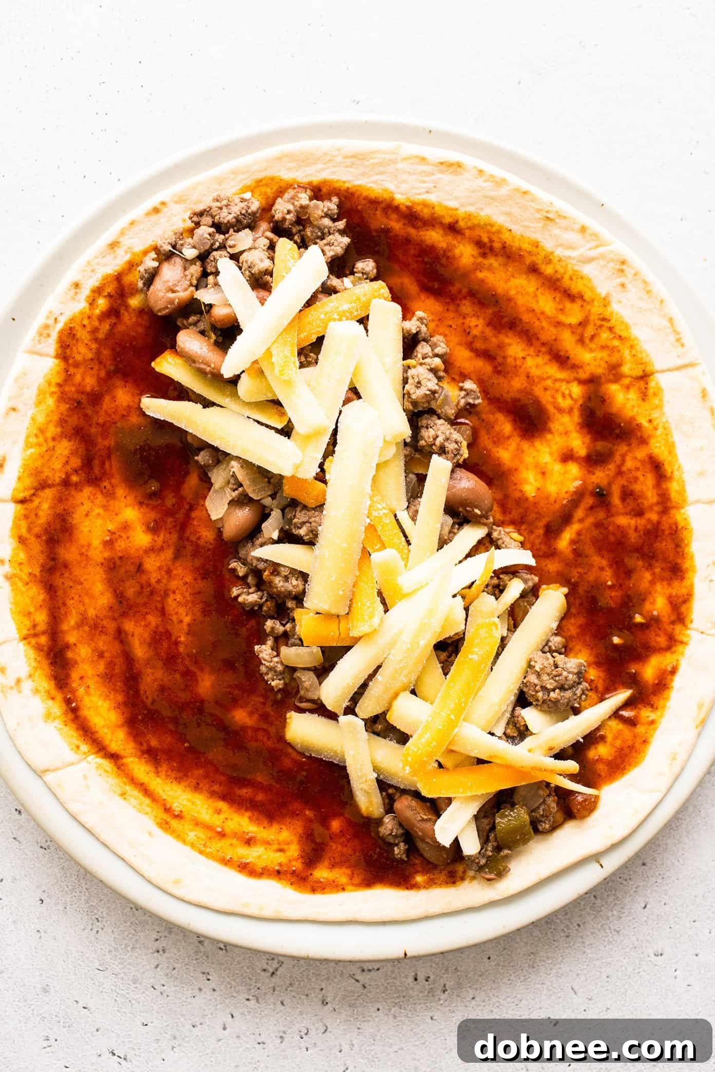 A close-up view of spreading the savory beef enchilada filling onto a tortilla, along with cheese and a drizzle of red enchilada sauce, demonstrating the assembly process.