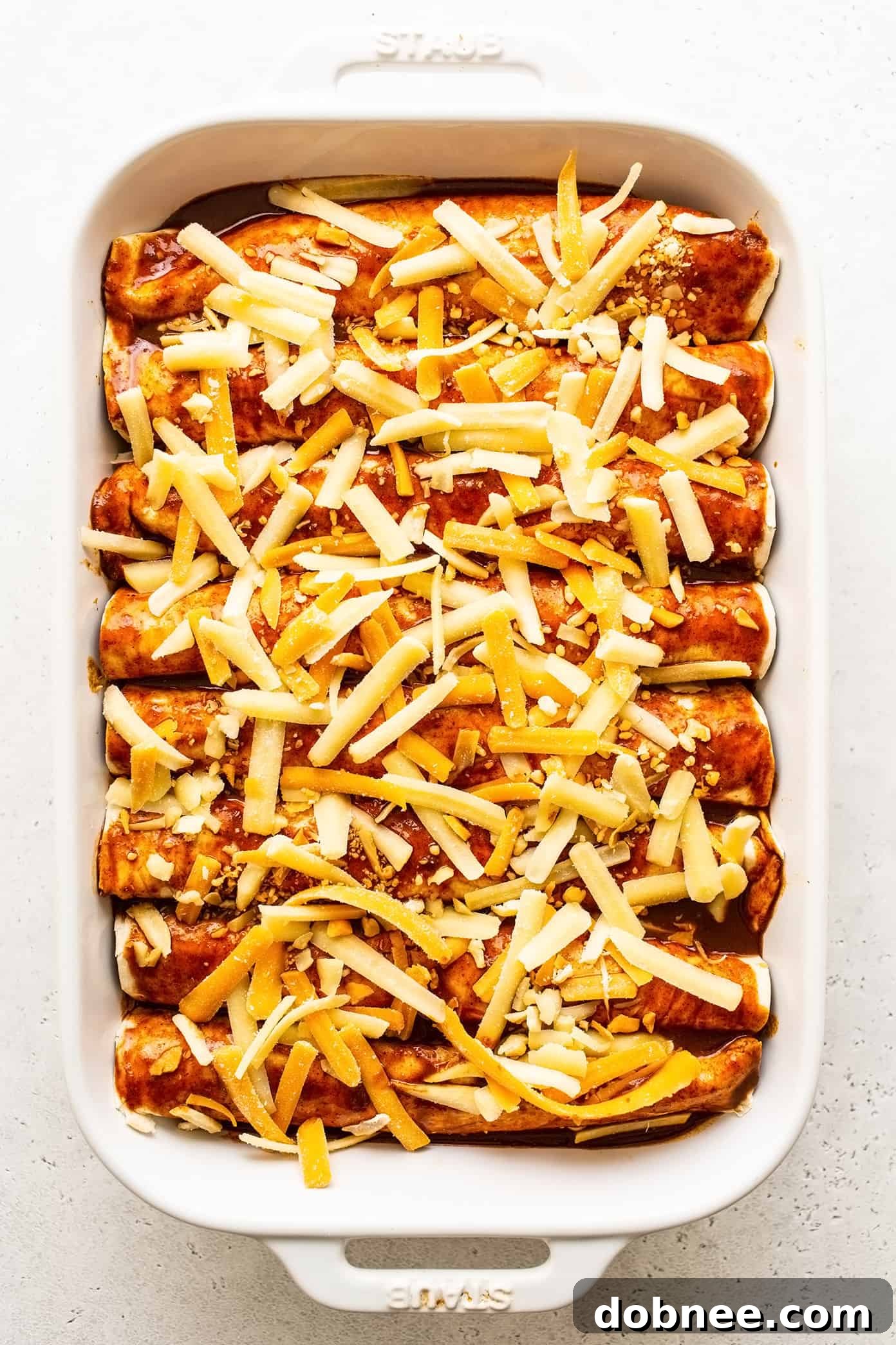 Uncooked beef enchiladas perfectly arranged in a 9x13 inch baking dish, generously topped with remaining red enchilada sauce and a final layer of shredded cheese, ready to be baked to golden perfection.