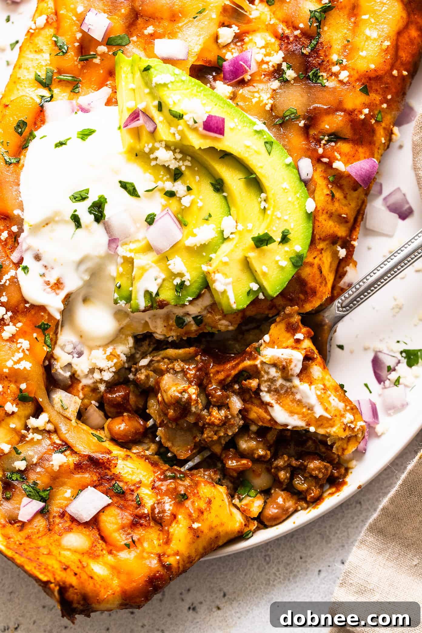 A beautifully baked beef enchilada, showcasing the rich filling and melted cheese, garnished with a vibrant slice of avocado and a dollop of sour cream for a refreshing contrast.