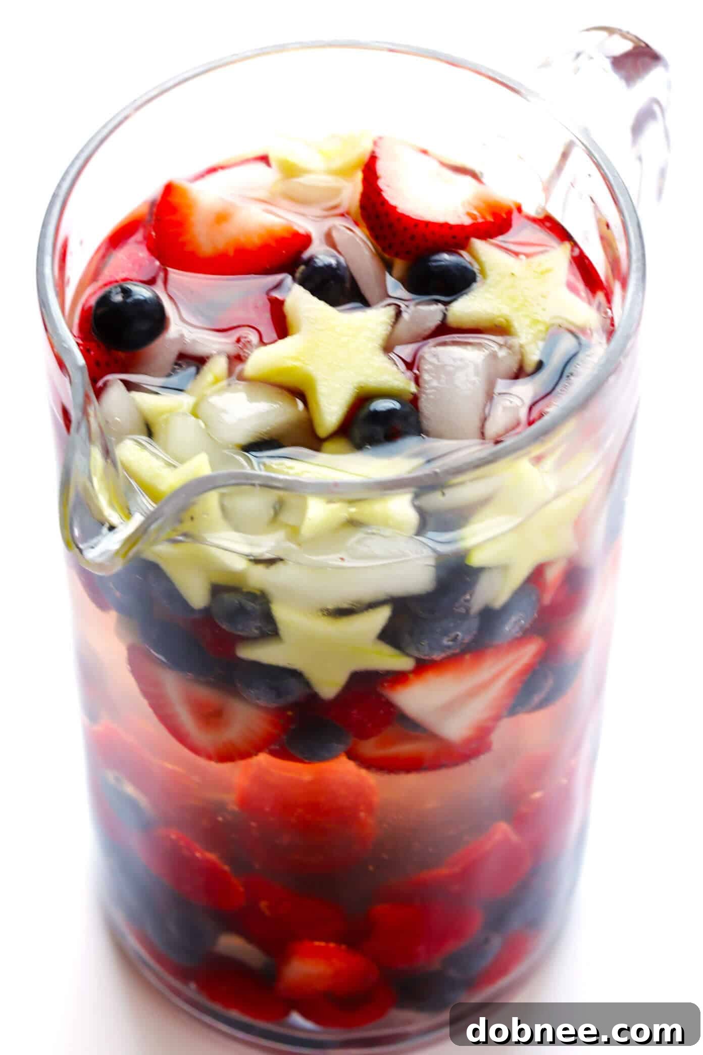 A pitcher brimming with Red, White, and Blue Sparkling Sangria, ready to be served over ice. Features strawberries, blueberries, and star-shaped apples for a festive holiday drink.