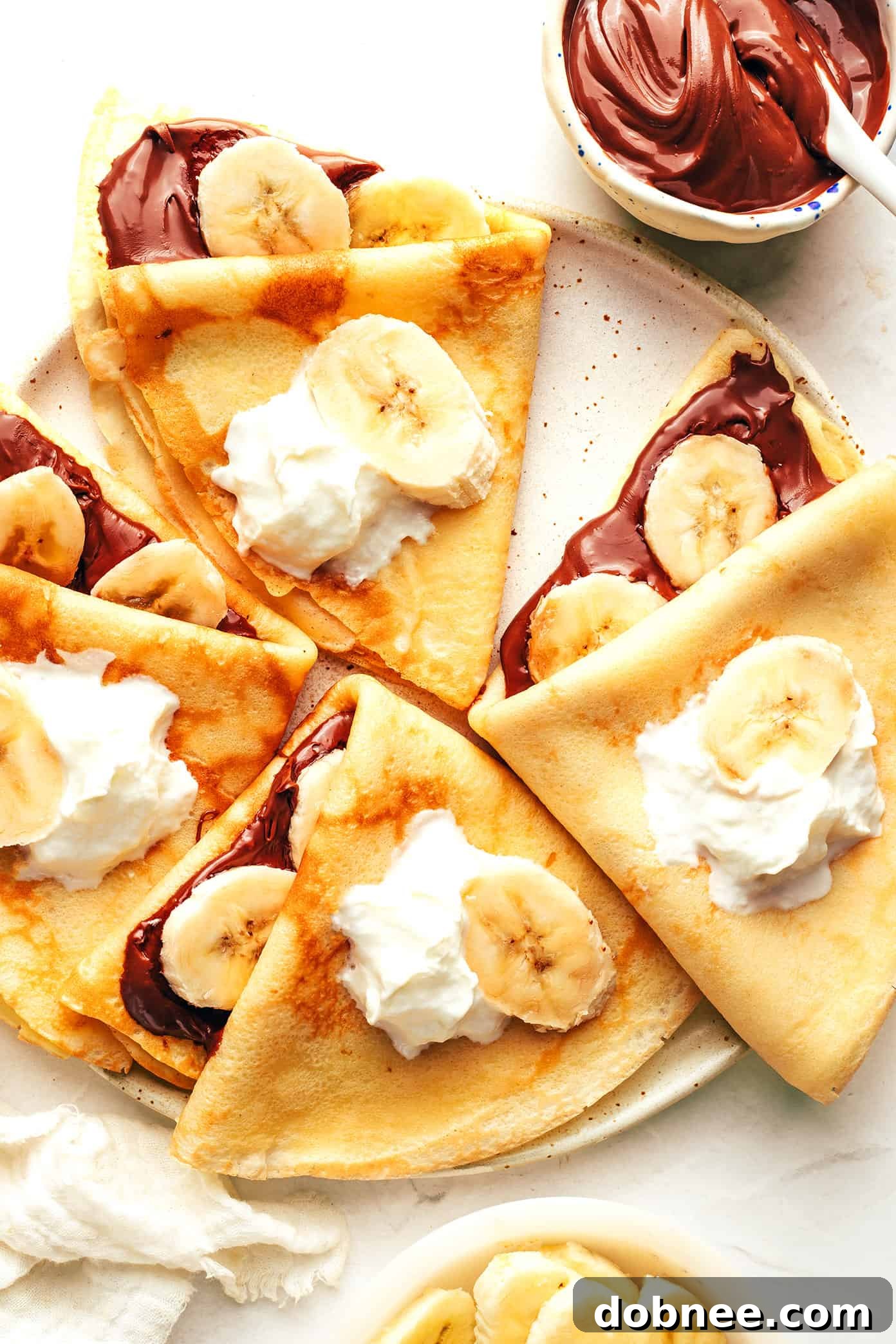 Two crepes folded in half, filled with sliced bananas and drizzled with Nutella, garnished with a mint sprig.