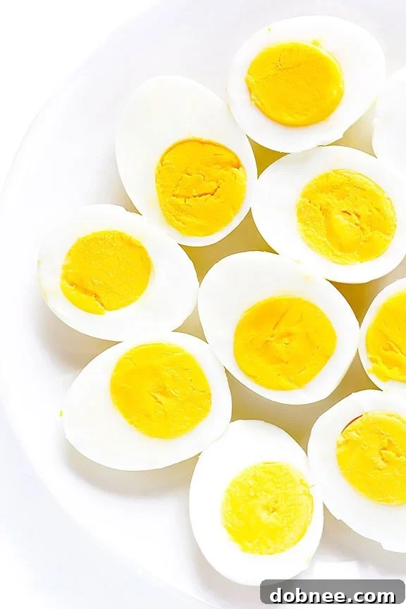 A beautifully peeled hard-boiled egg cut in half, showcasing a perfectly firm yellow yolk, sitting on a wooden board next to whole hard-boiled eggs. This image demonstrates the ideal result of making perfect hard-boiled eggs.