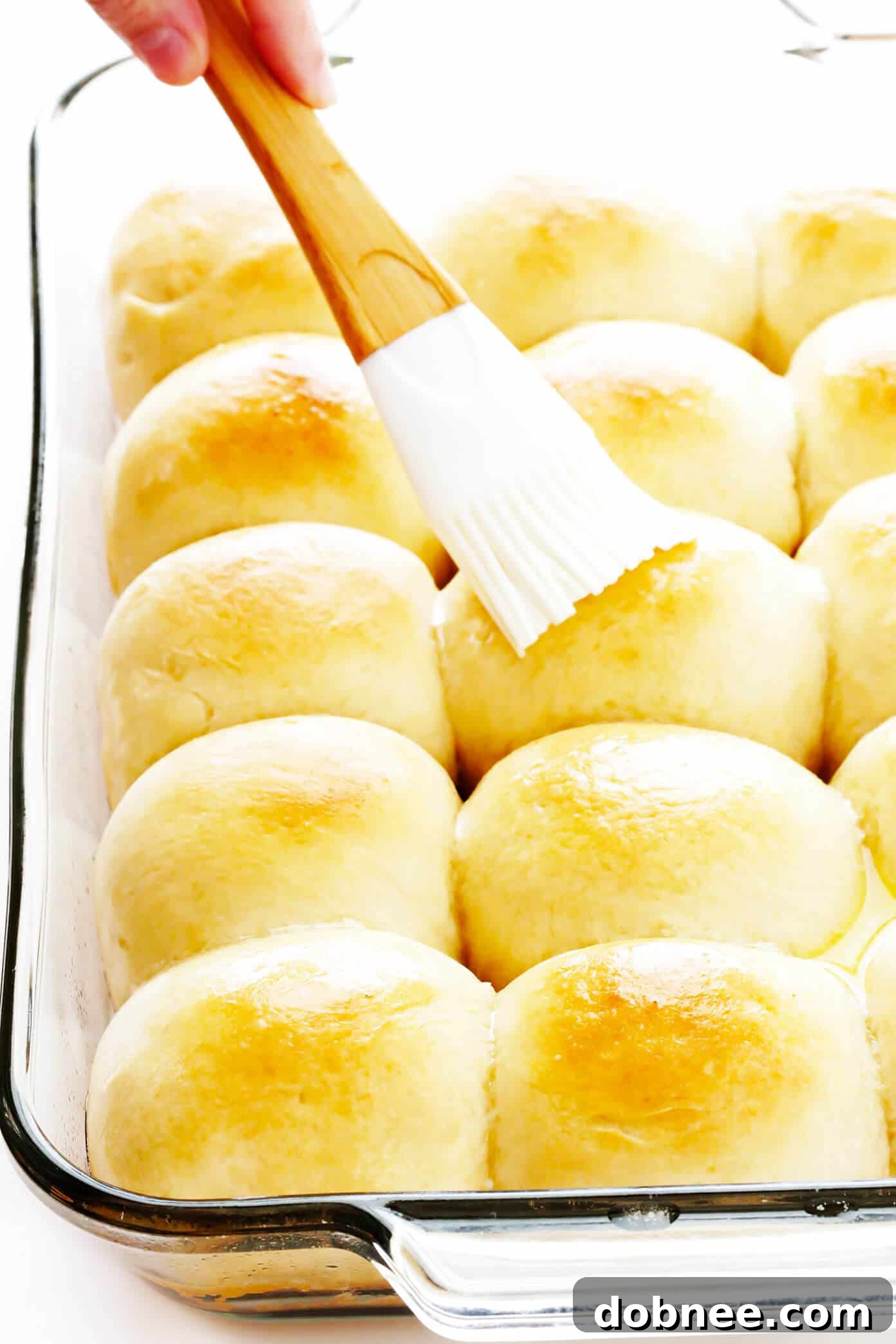 A hand brushing melted butter over golden brown dinner rolls fresh out of the oven in a baking dish.