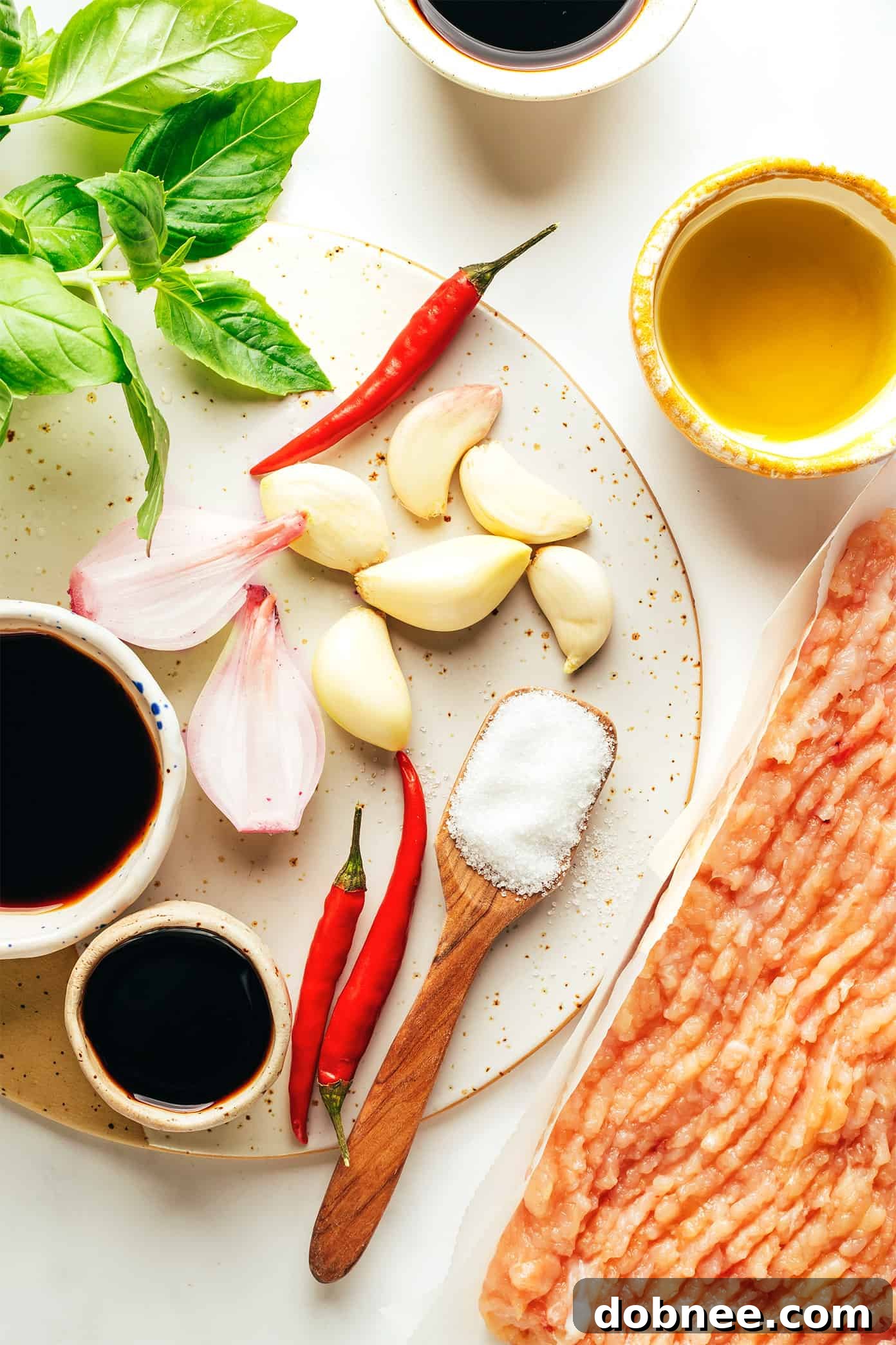 Ingredients for Thai Basil Chicken laid out on a cutting board, including fresh holy basil leaves, minced garlic, red chili peppers, chopped shallots, and ground chicken, indicating preparation for a delicious stir-fry.