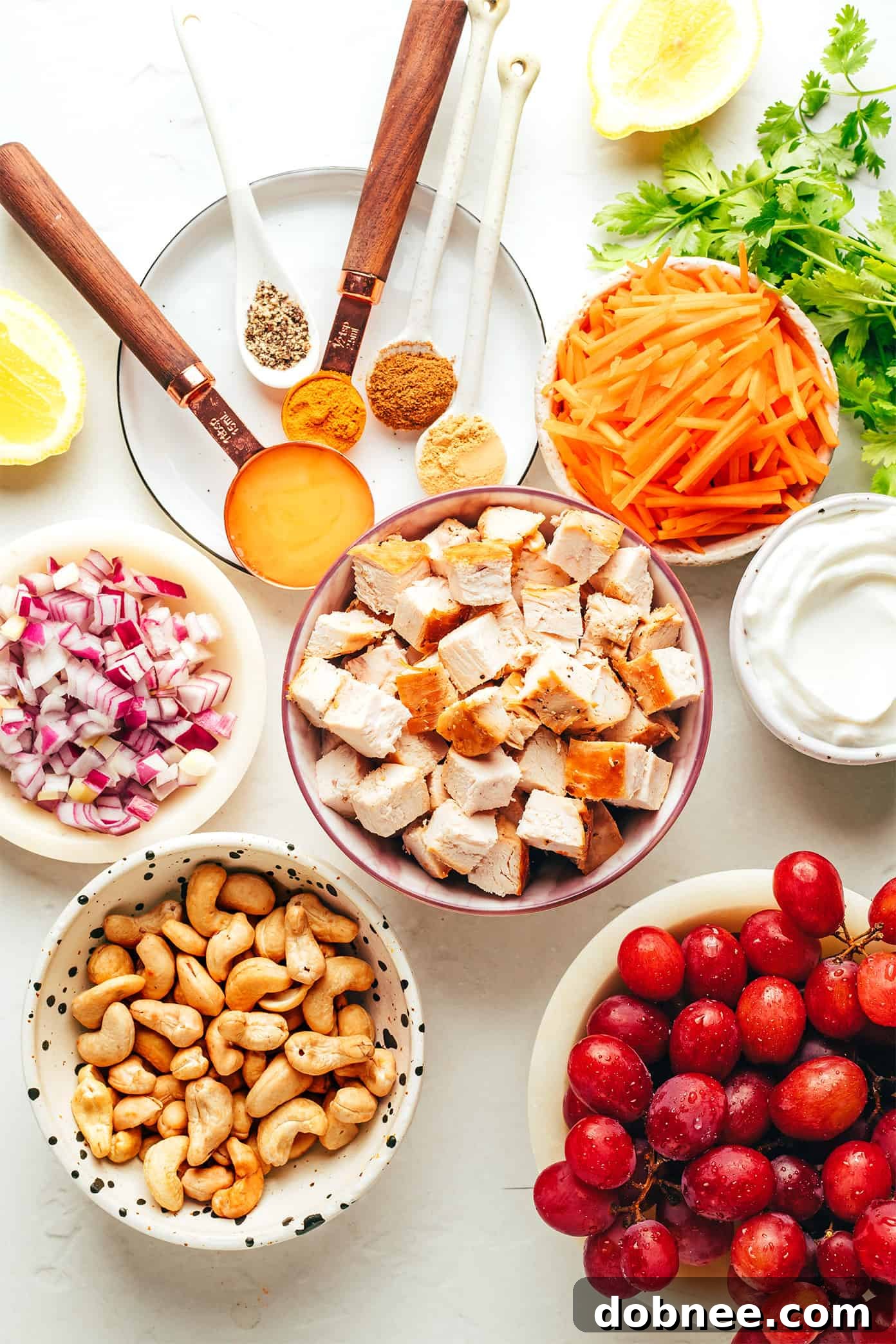 Assortment of fresh ingredients for healthy curry chicken salad, including diced chicken, grapes, cilantro, carrots, cashews, and red onion.