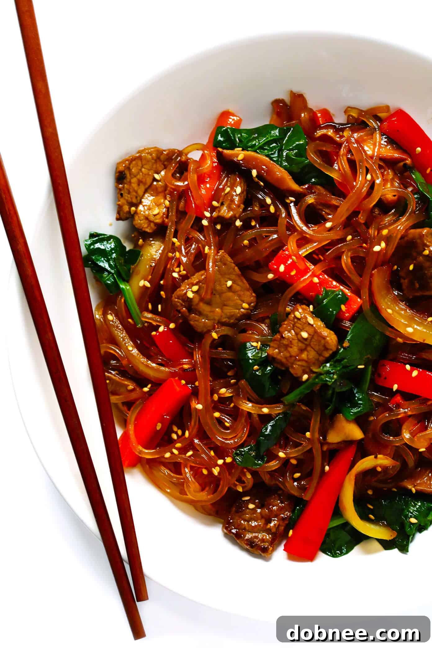 A vibrant Korean Japchae stir-fry with colorful vegetables, beef, and translucent sweet potato glass noodles, garnished with sesame seeds.