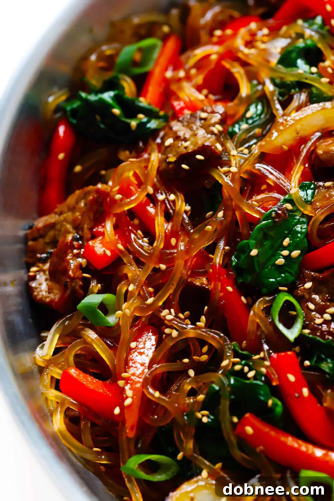 A beautifully plated bowl of Japchae with beef, colorful vegetables, and glass noodles, garnished with sesame seeds and green onions.