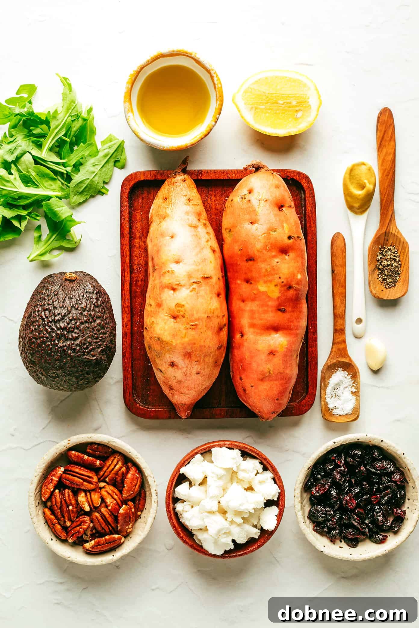 A vibrant display of fresh ingredients for the Roasted Sweet Potato and Arugula Salad, including sweet potatoes, arugula, avocado, goat cheese, pecans, dried cranberries, and lemon.