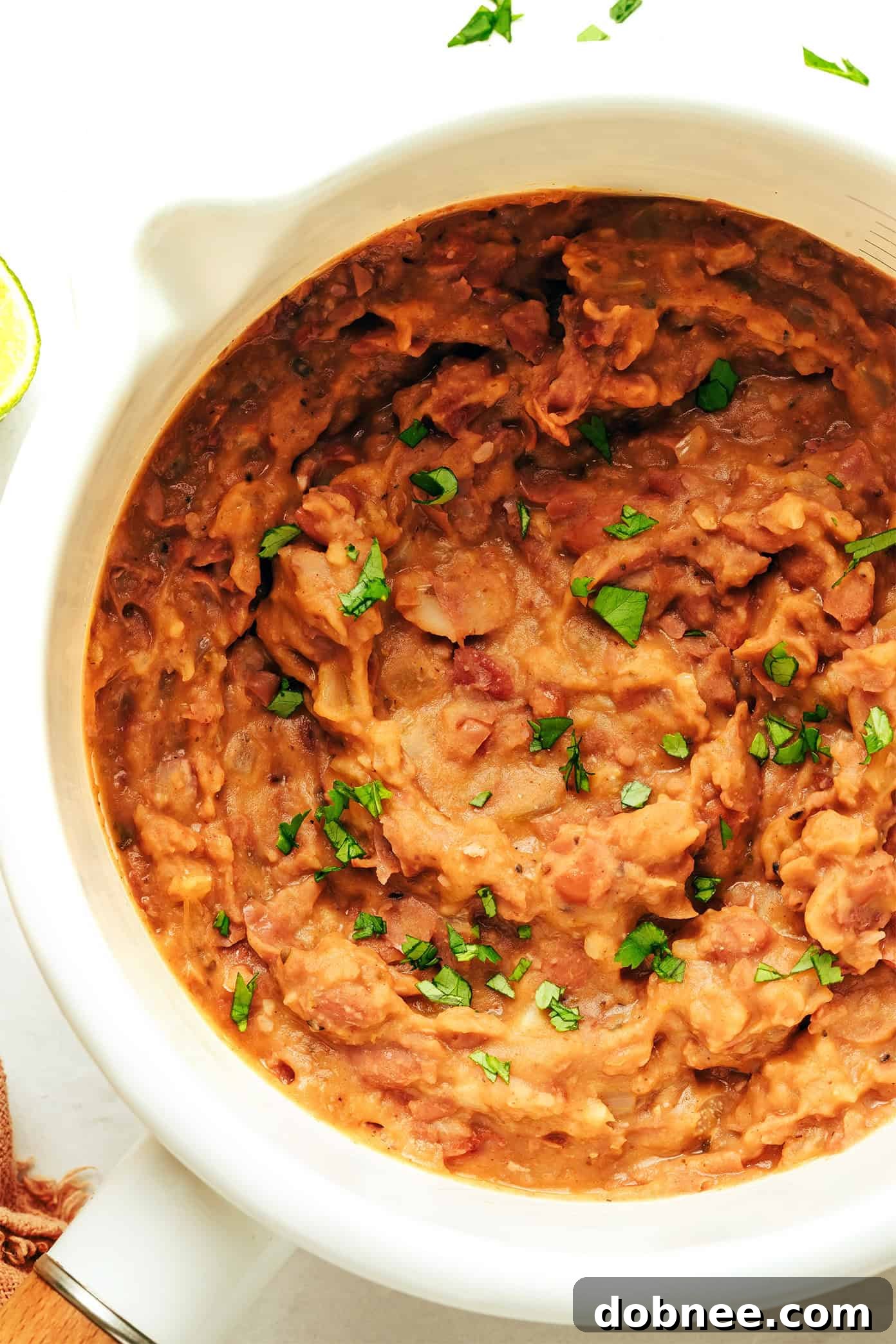 A serving of homemade refried beans topped with crumbled cotija cheese and fresh cilantro, alongside tortilla chips and a lime wedge.