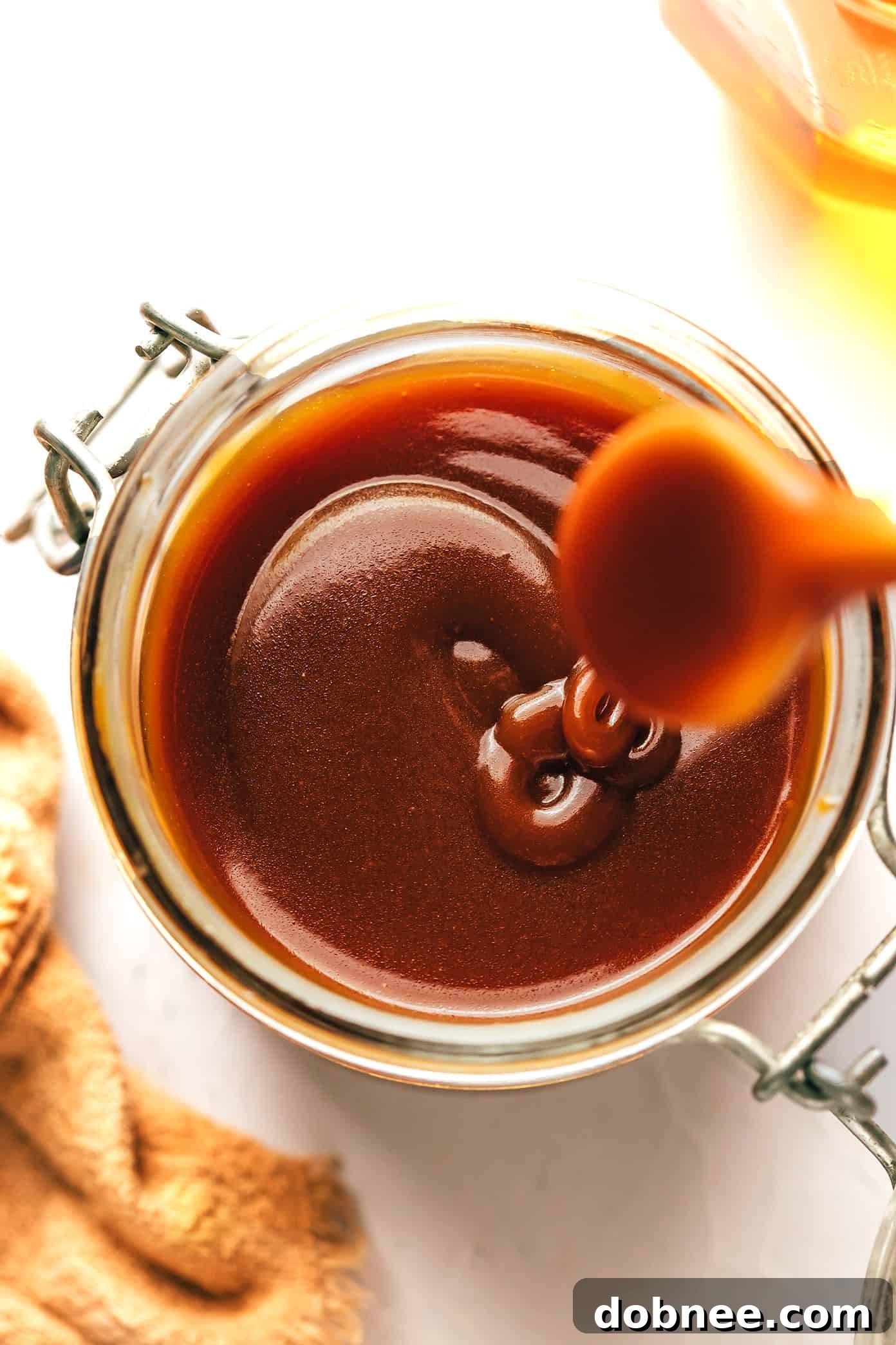 A jar of homemade bourbon caramel sauce, with a spoon drizzling the sauce onto a serving of ice cream, showcasing its texture.
