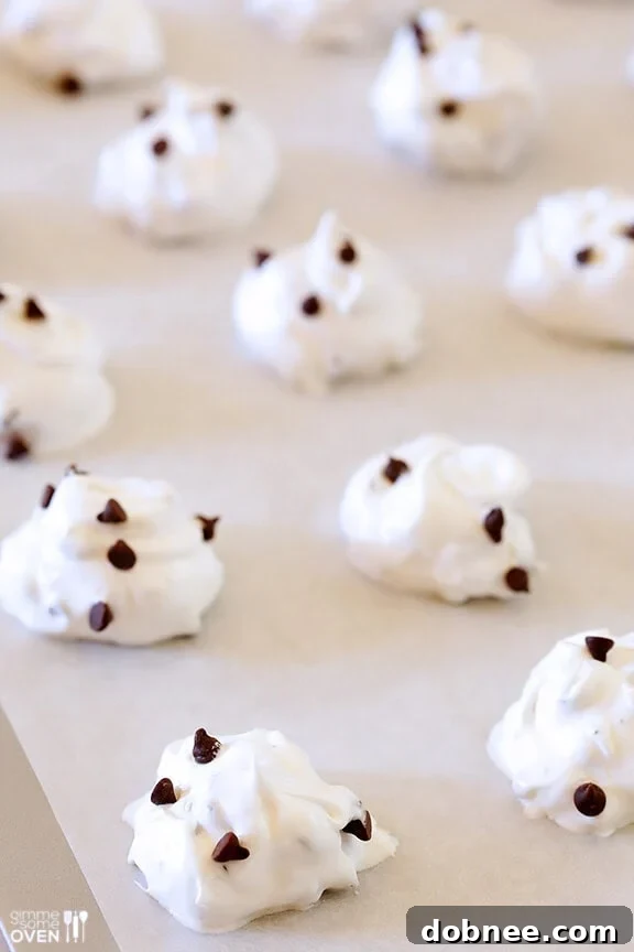 Unbaked chocolate chip meringue cookie dollops neatly arranged on a parchment-lined baking sheet, ready for the oven.