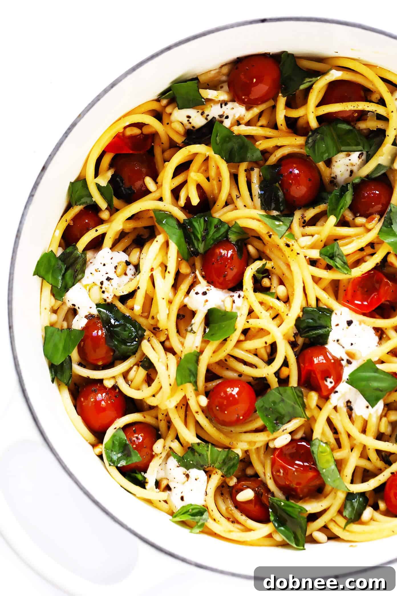 A large pot of bucatini pasta mixed with vibrant burst cherry tomatoes, melting burrata cheese, fresh basil leaves, and crunchy pine nuts, ready to serve