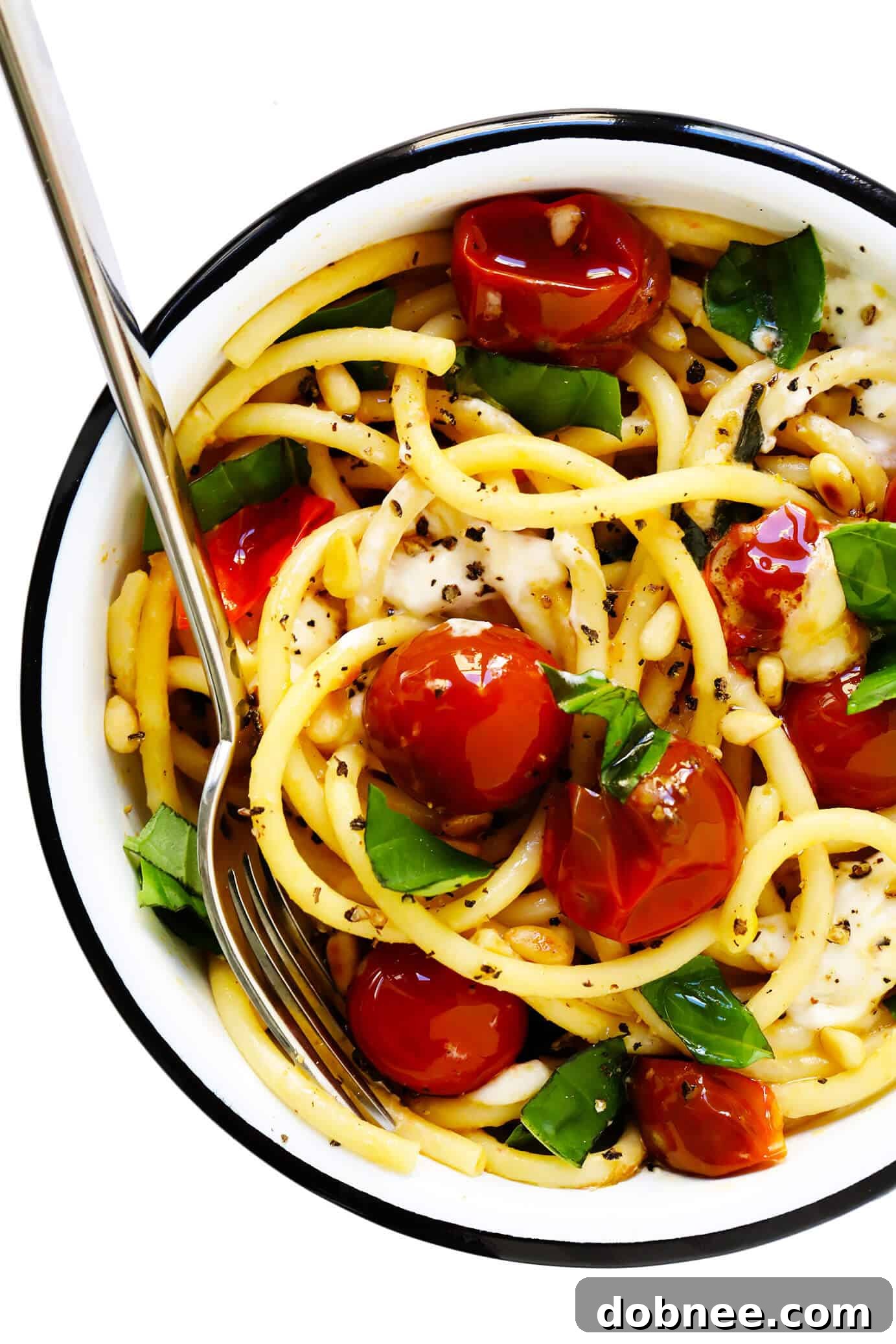 A beautifully presented serving bowl of garlicky pasta, featuring vibrant burst cherry tomatoes, melted burrata, fresh basil, and a sprinkle of pine nuts