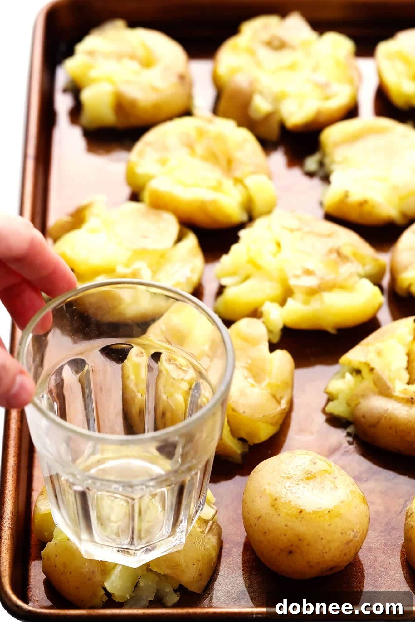 A hand gently smashing a boiled potato on a baking sheet using the bottom of a drinking glass, illustrating a key step in making smashed potatoes.