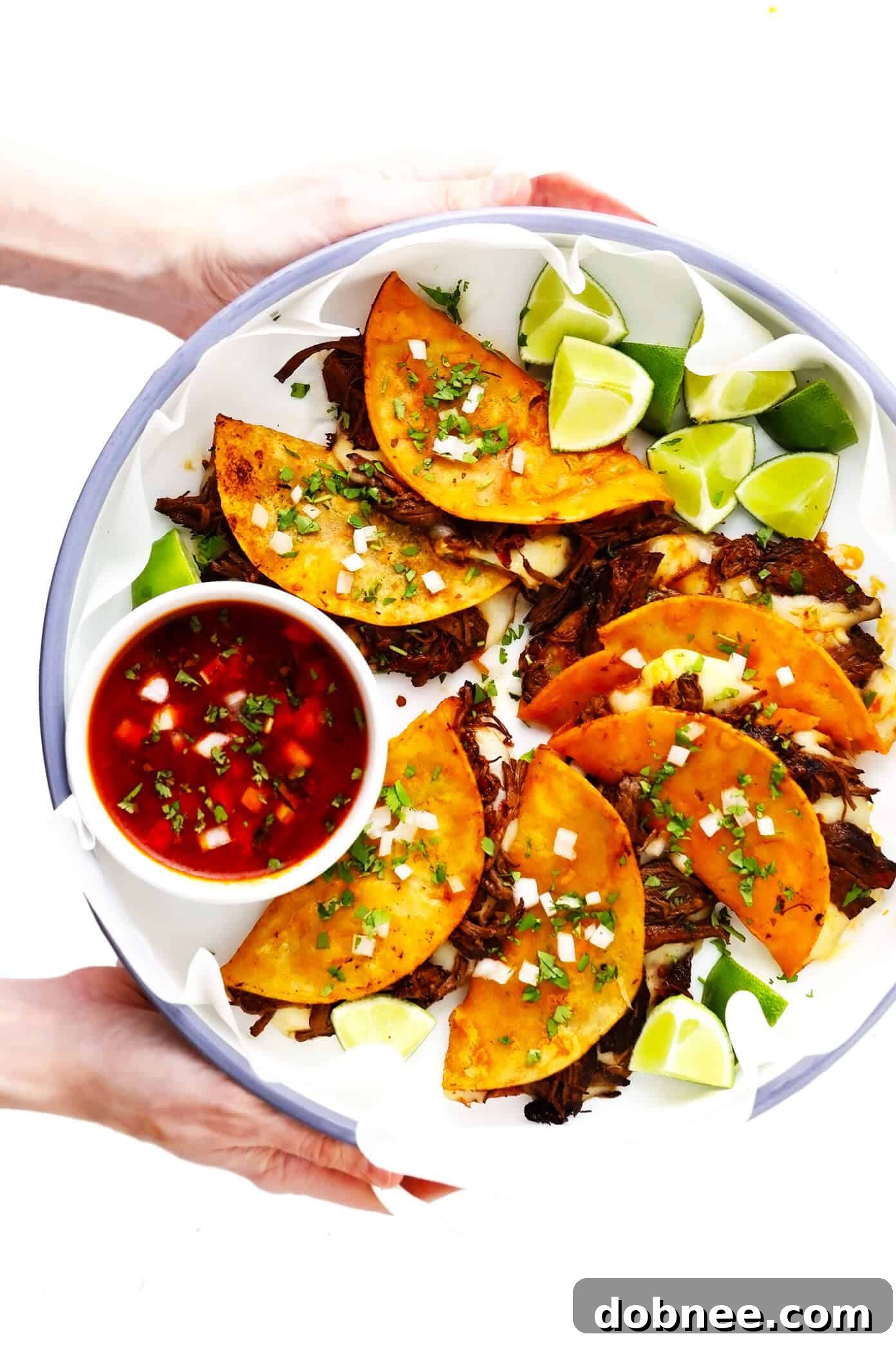 Plated Birria Quesatacos with a bowl of consommé for dipping and fresh lime wedges.