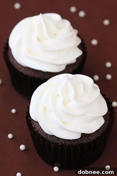 Close-up shot of two freshly baked White Chocolate Mocha Cupcakes, showcasing the fluffy white chocolate whipped cream frosting and a sprinkle of cocoa powder.