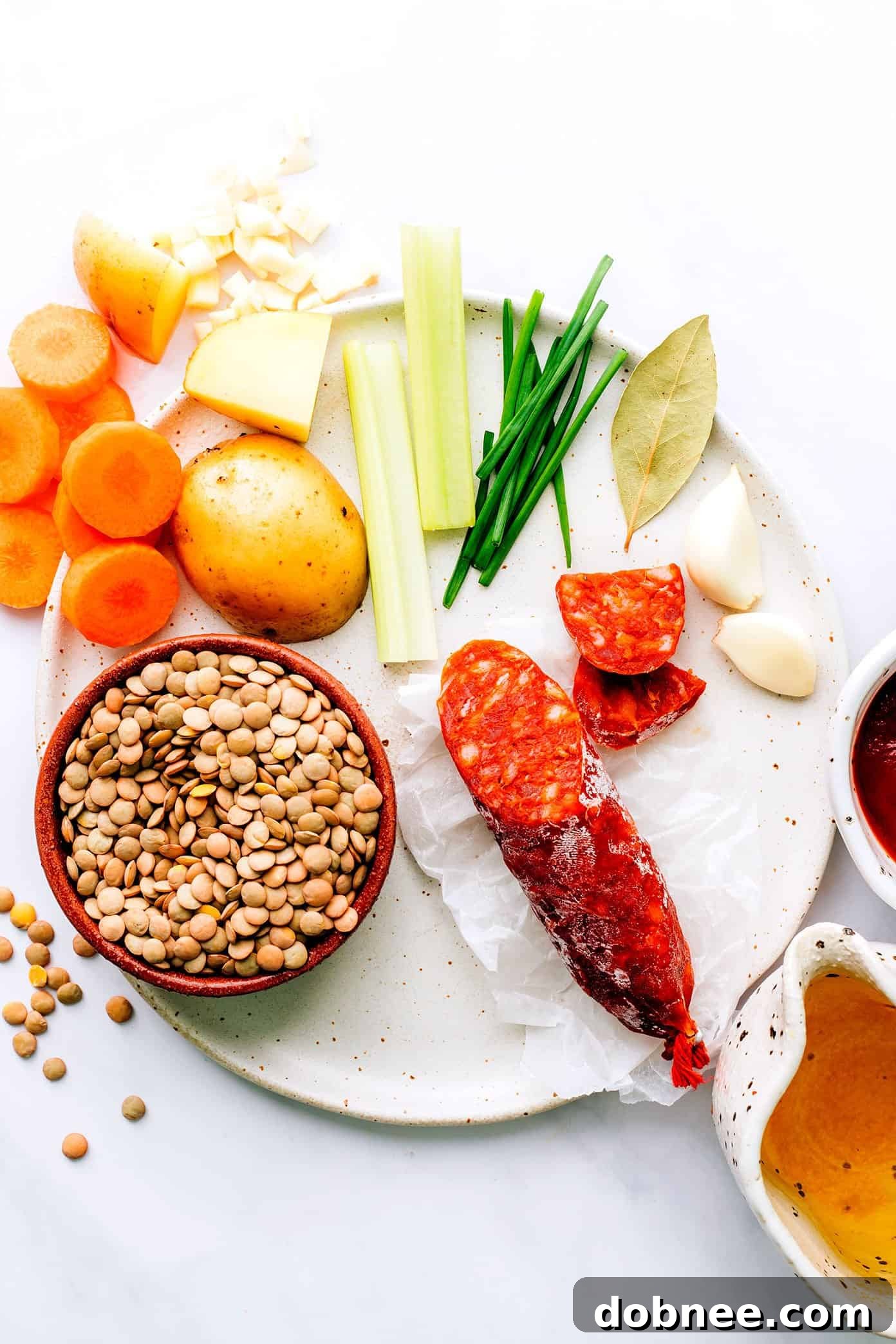 Individual ingredients for Spanish Chorizo Lentil Stew laid out, including carrots, potato, garlic, celery, chives, bay leaf, tomato paste, and broth