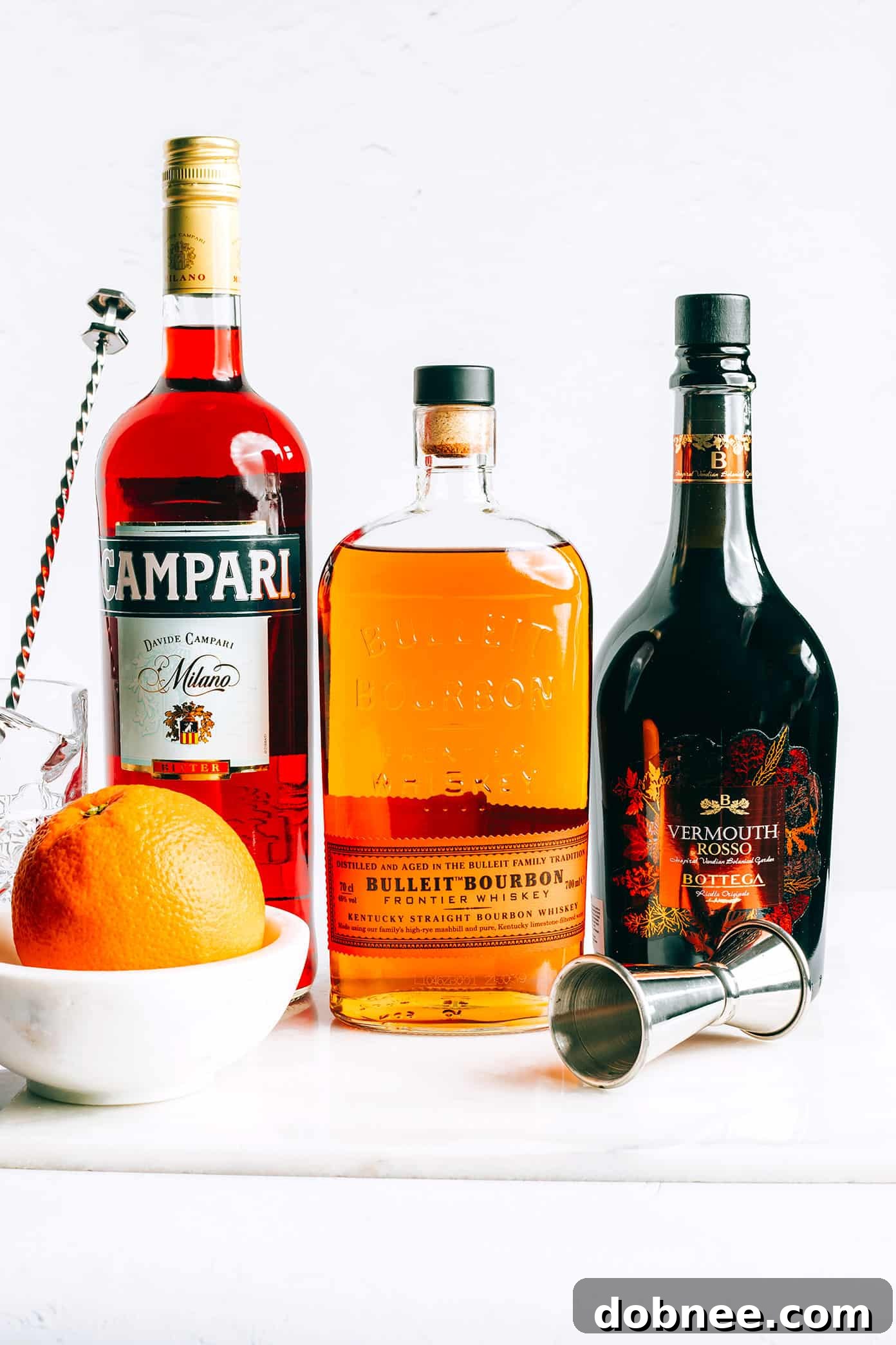 Essential Boulevardier ingredients laid out on a wooden surface: a bottle of Campari, a bottle of bourbon, sweet vermouth, and fresh orange peel.