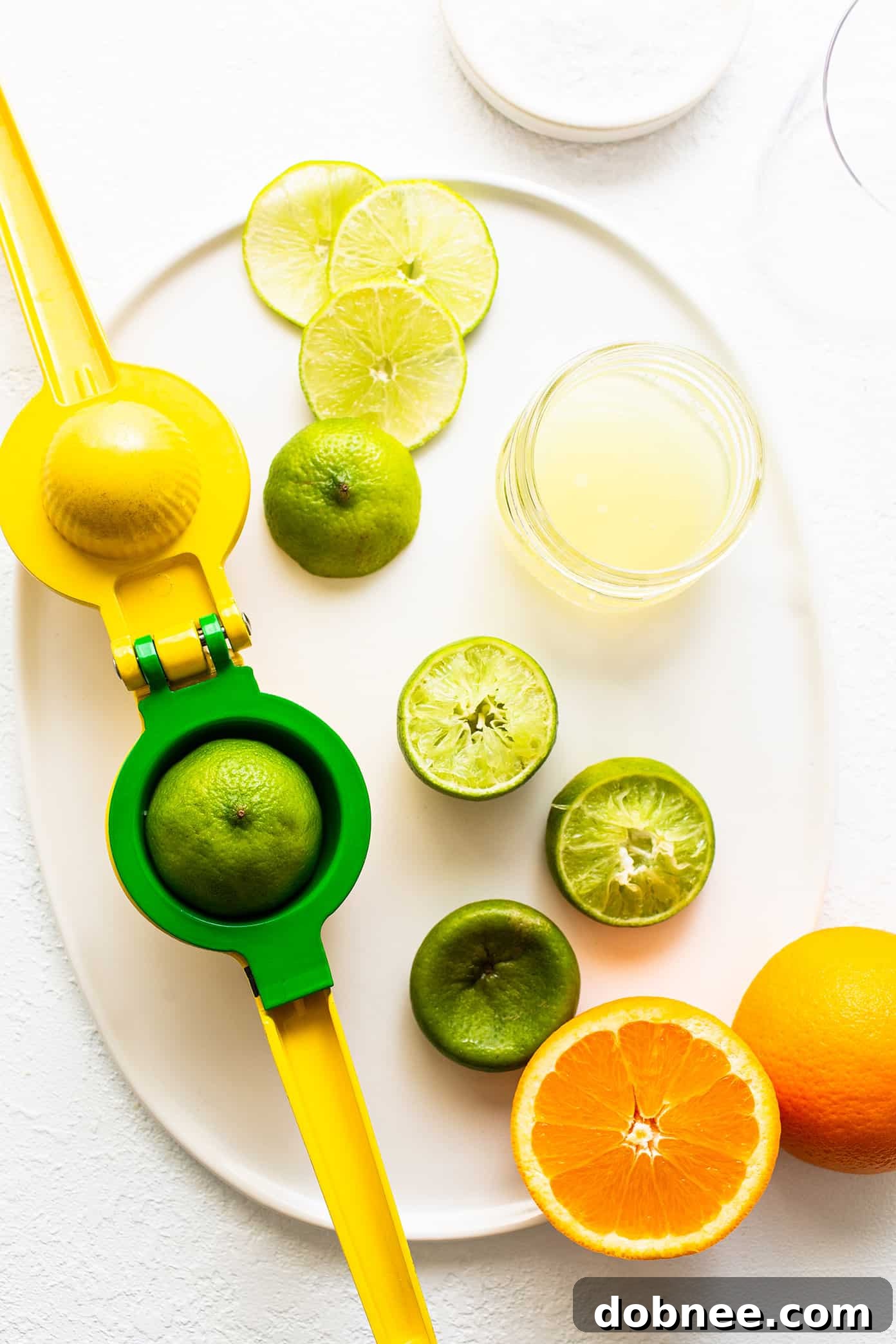 Fresh ingredients laid out on a plate for making a Virgin Margarita Mocktail: limes, an orange, and a bottle of agave nectar.