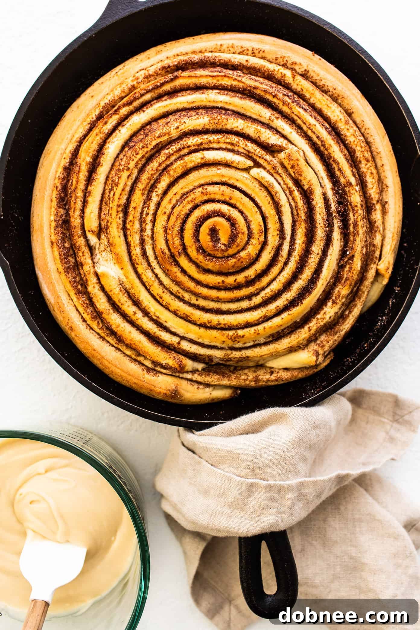 Giant Cinnamon Roll Ready for Icing An unfrosted giant cinnamon roll nestled in a cast iron skillet, with a bowl of creamy cream cheese frosting placed beside it, ready for application.