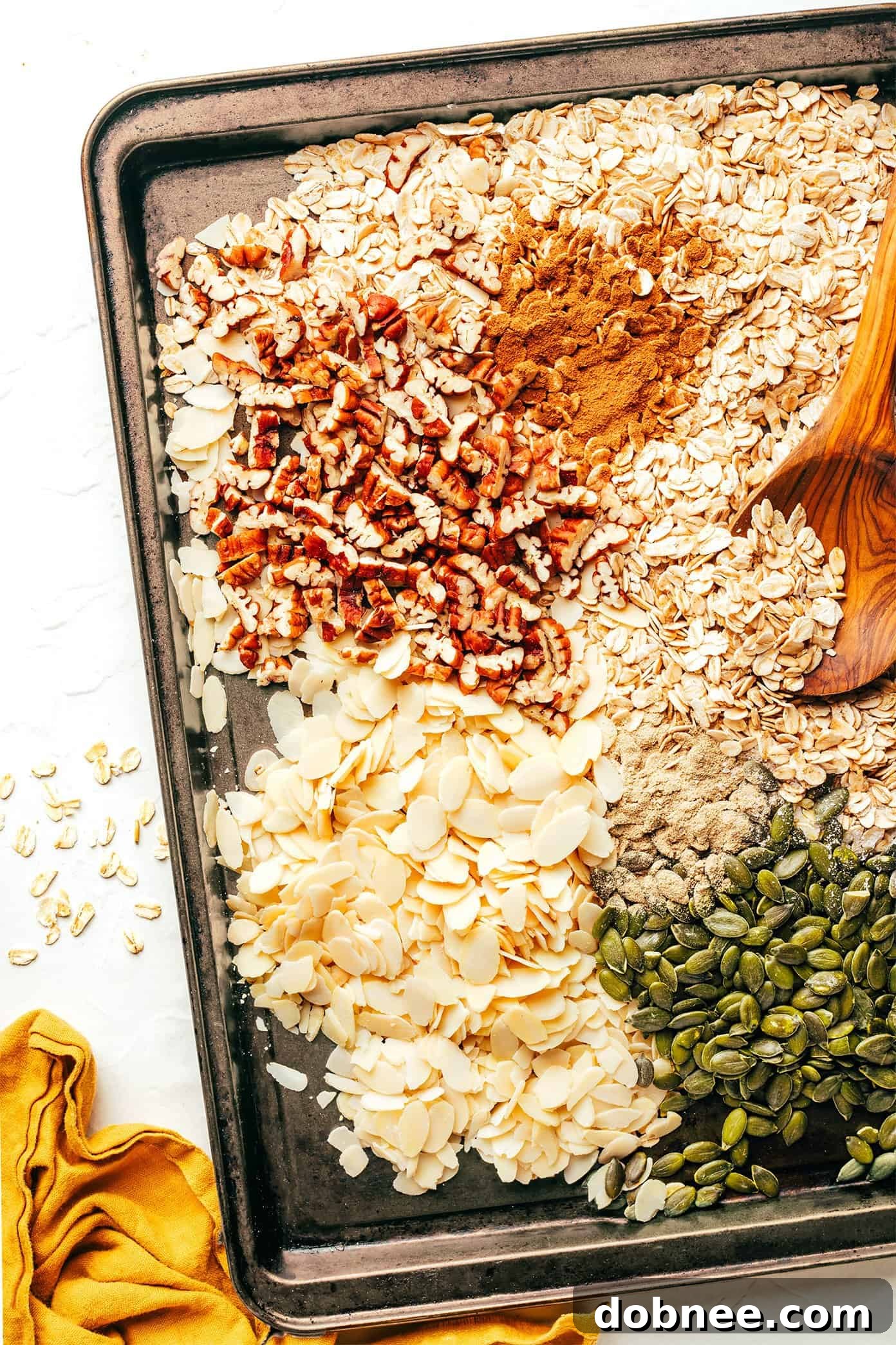 Raw oats, nuts, and spices spread evenly on a baking sheet, ready for toasting.