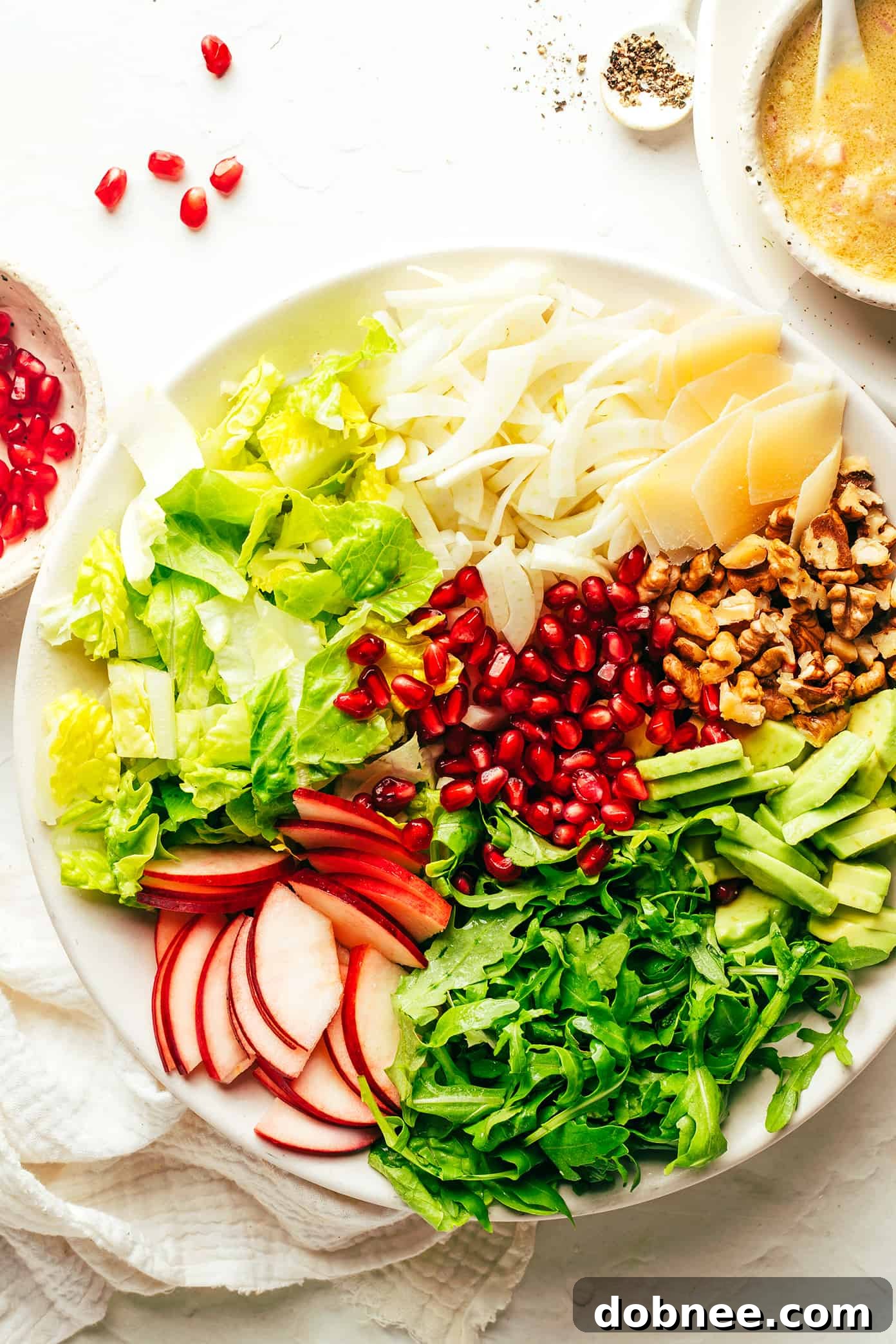 A flat lay image displaying all the fresh and vibrant ingredients needed for the Holiday Green Salad, including pomegranate, apple, arugula, avocado, walnuts, Parmesan, fennel, and components for shallot vinaigrette.