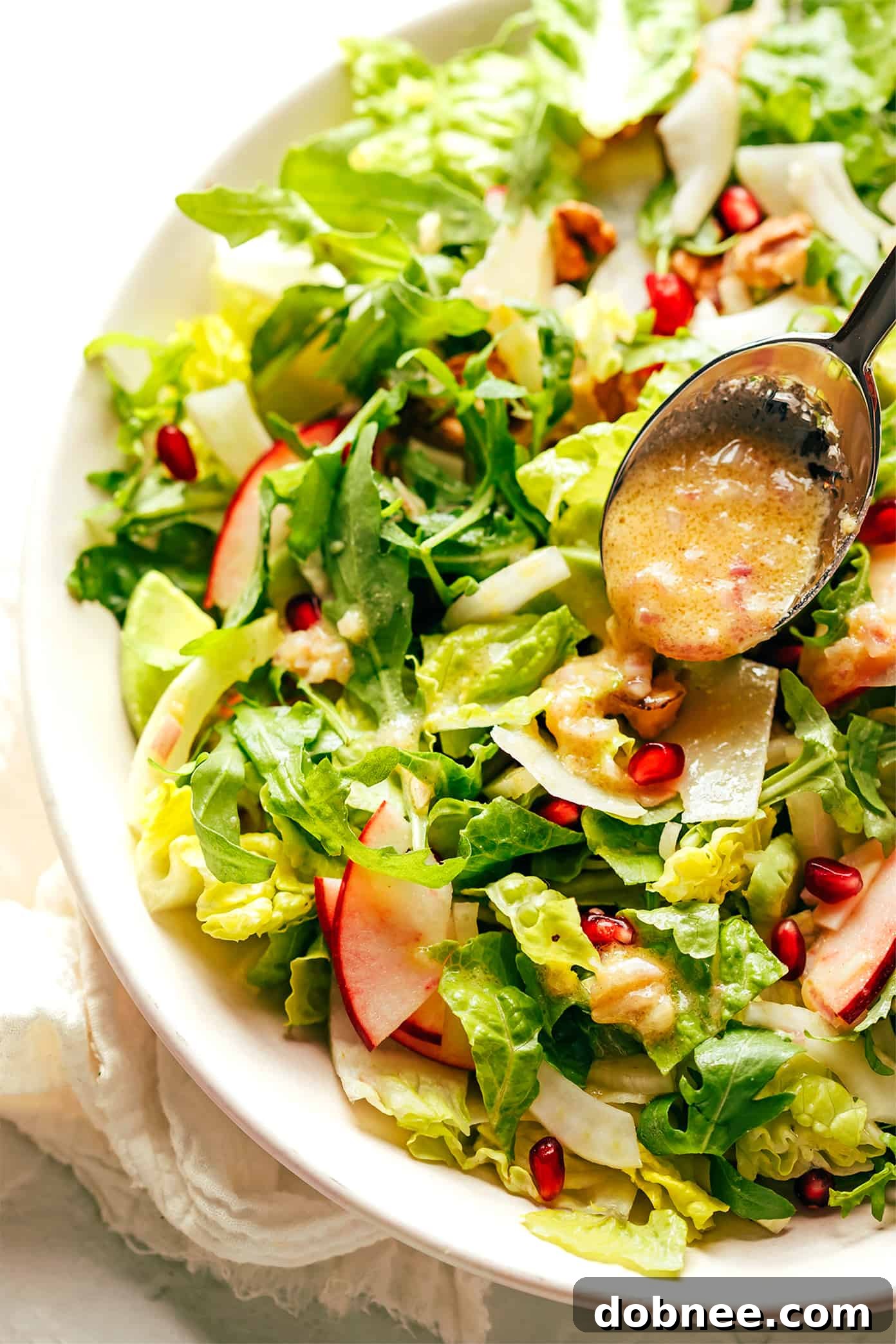 Close-up shot of a large mixing bowl, showing a Holiday Green Salad being drizzled with shallot vinaigrette and gently tossed to combine, highlighting the vibrant colors of pomegranate, apple, and greens.