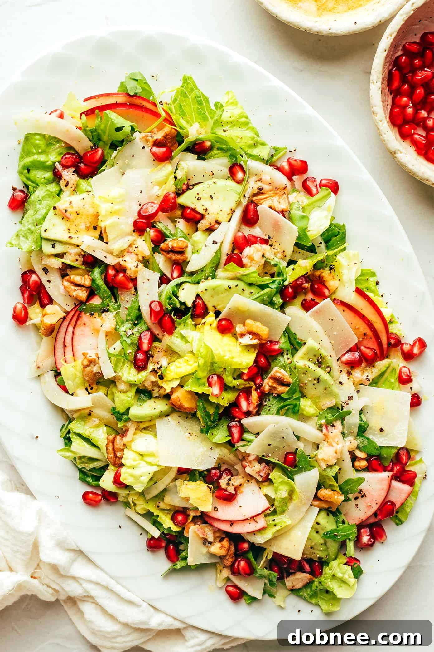 A festive Holiday Green Salad, richly adorned with pomegranate, apple, avocado, shaved Parmesan, fennel, and toasted walnuts, beautifully arranged on a serving platter, ready for a Thanksgiving or Christmas meal.
