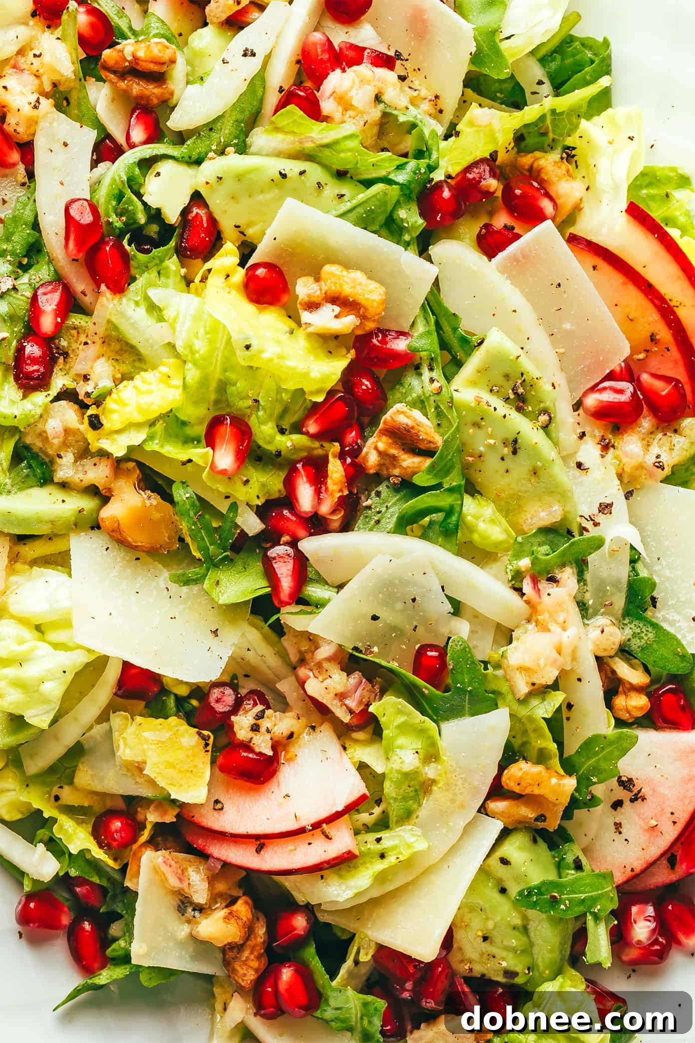 A mesmerizing close-up of the Christmas Pomegranate Salad, highlighting the intricate details of the pomegranate arils, thinly sliced apple, delicate greens, and shaved Parmesan, all glistening with vinaigrette.