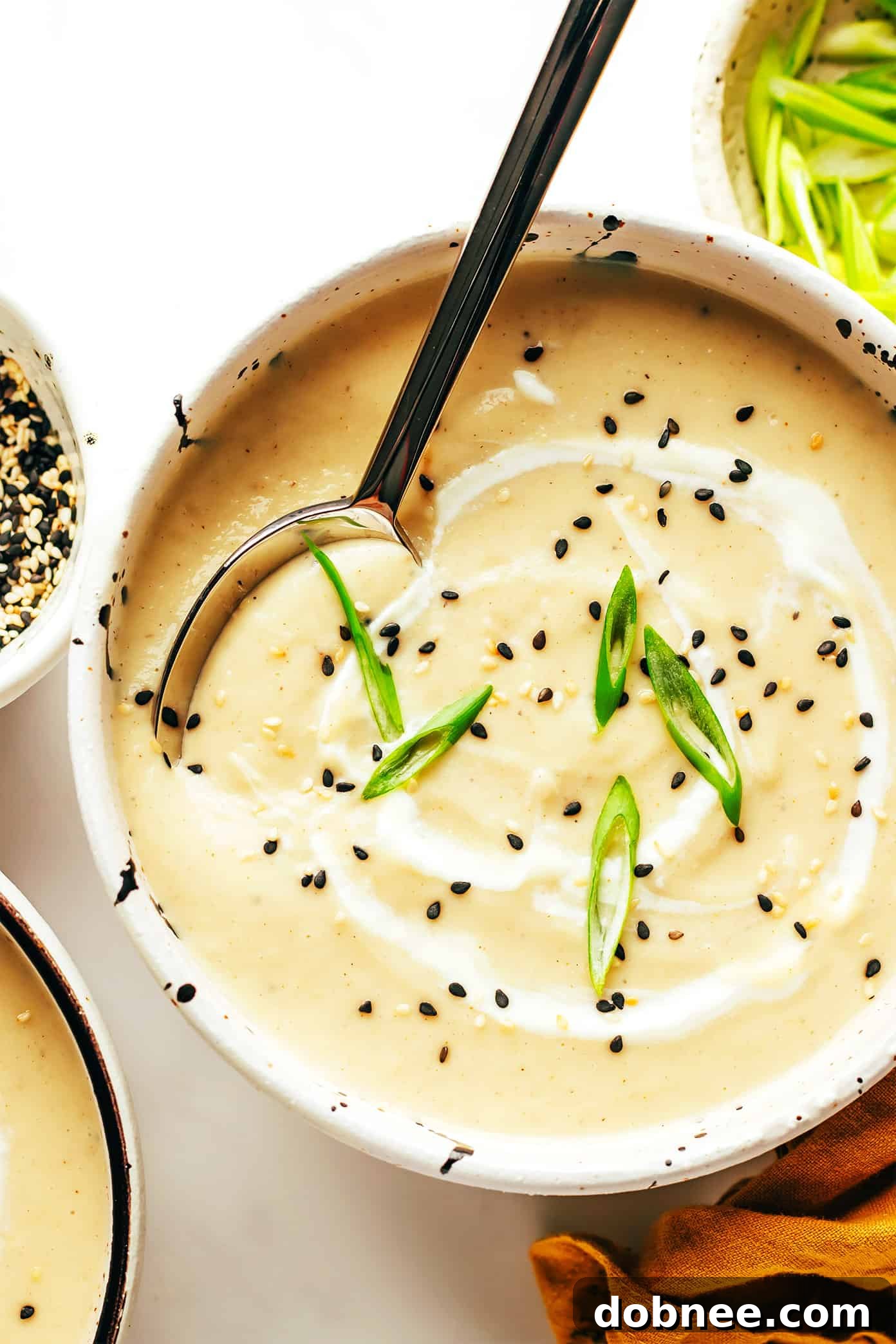 A bowl of creamy sesame ginger roasted cauliflower soup garnished with green onions and sesame seeds, with a spoon dipping in.