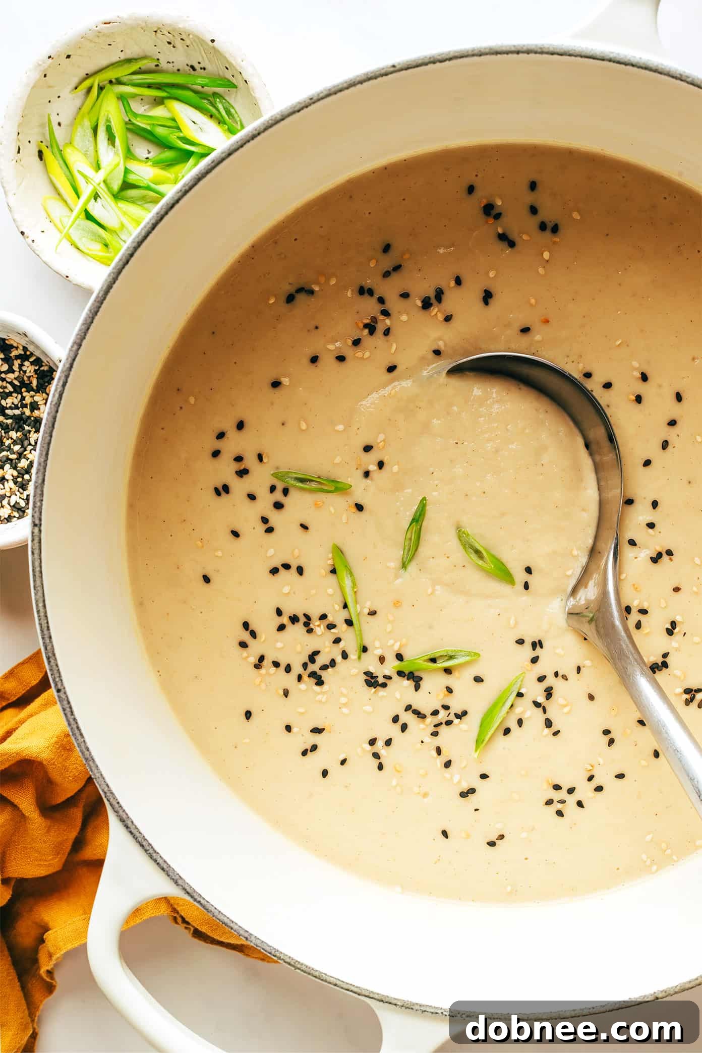 A close-up of a hand pouring creamy coconut milk into a bowl of sesame ginger roasted cauliflower soup, with green onions and sesame seeds on top.