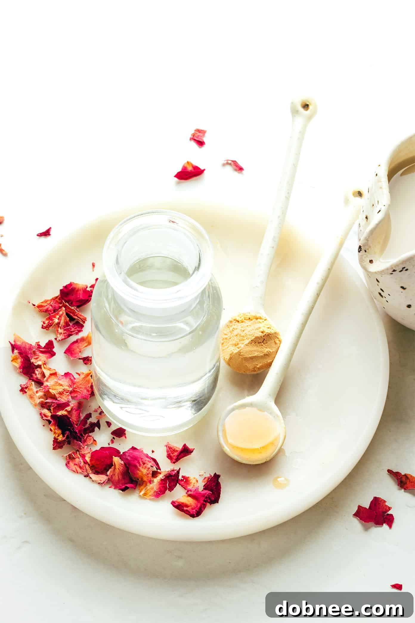 Velvet Rose Latte 3 Ingredients for a homemade rose latte laid out on a rustic surface: milk, rose water, maple syrup, vanilla extract, ground ginger, and dried rose petals.