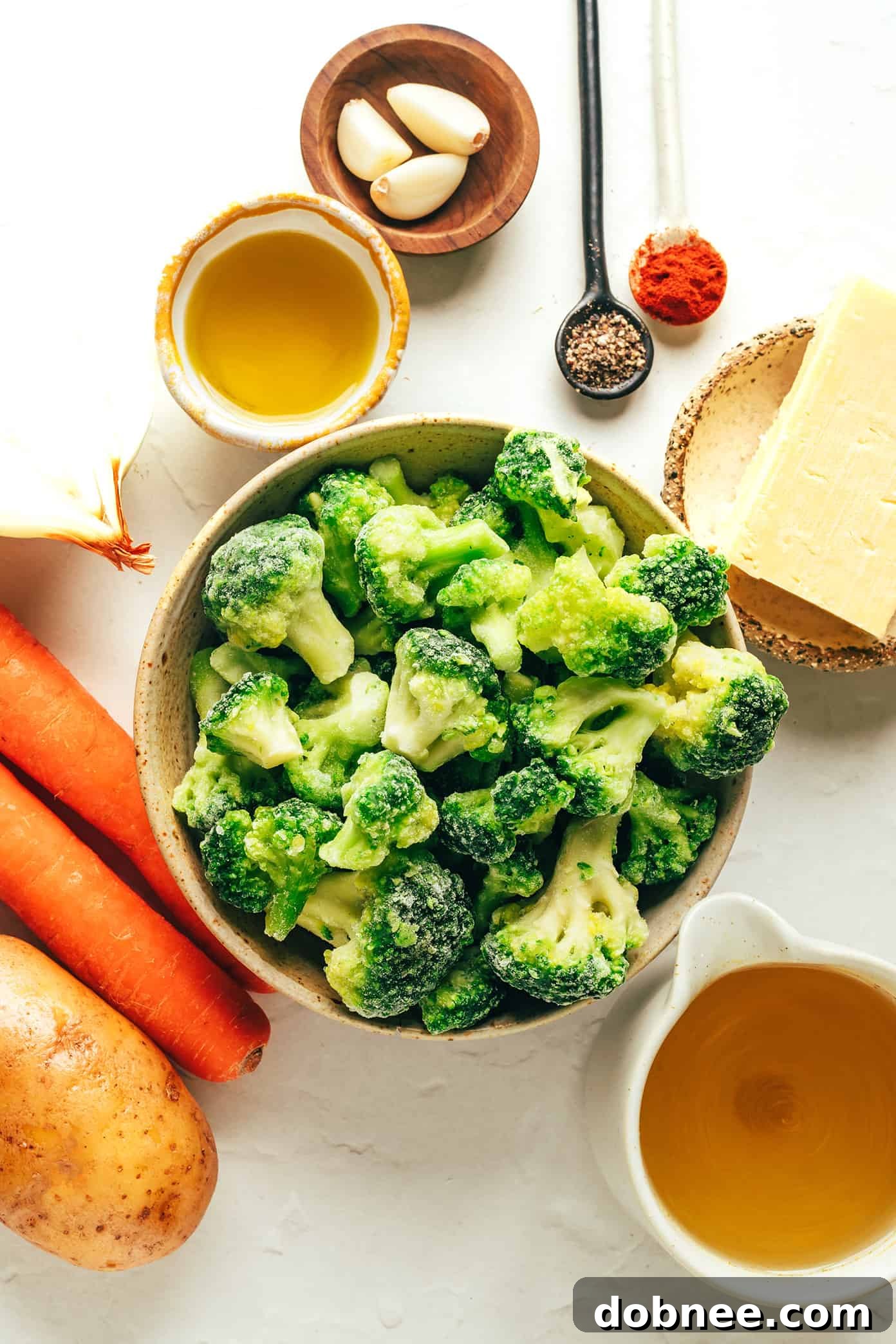 Fresh ingredients laid out for making broccoli cheese soup: broccoli florets, potatoes, carrots, onions, garlic, and a block of cheddar cheese.
