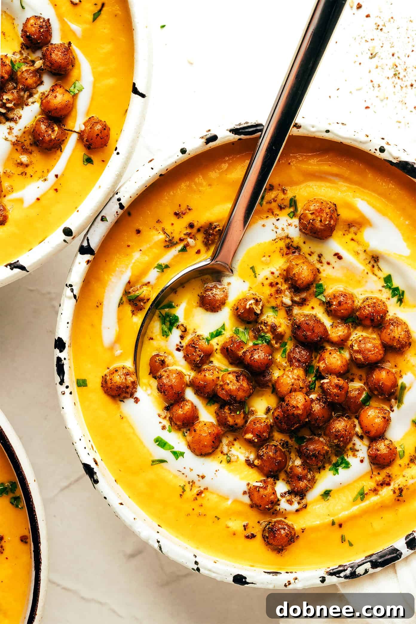 A vibrant bowl of roasted carrot soup with a swirl of cream and a sprinkle of crispy za'atar chickpeas, served with fresh herbs.