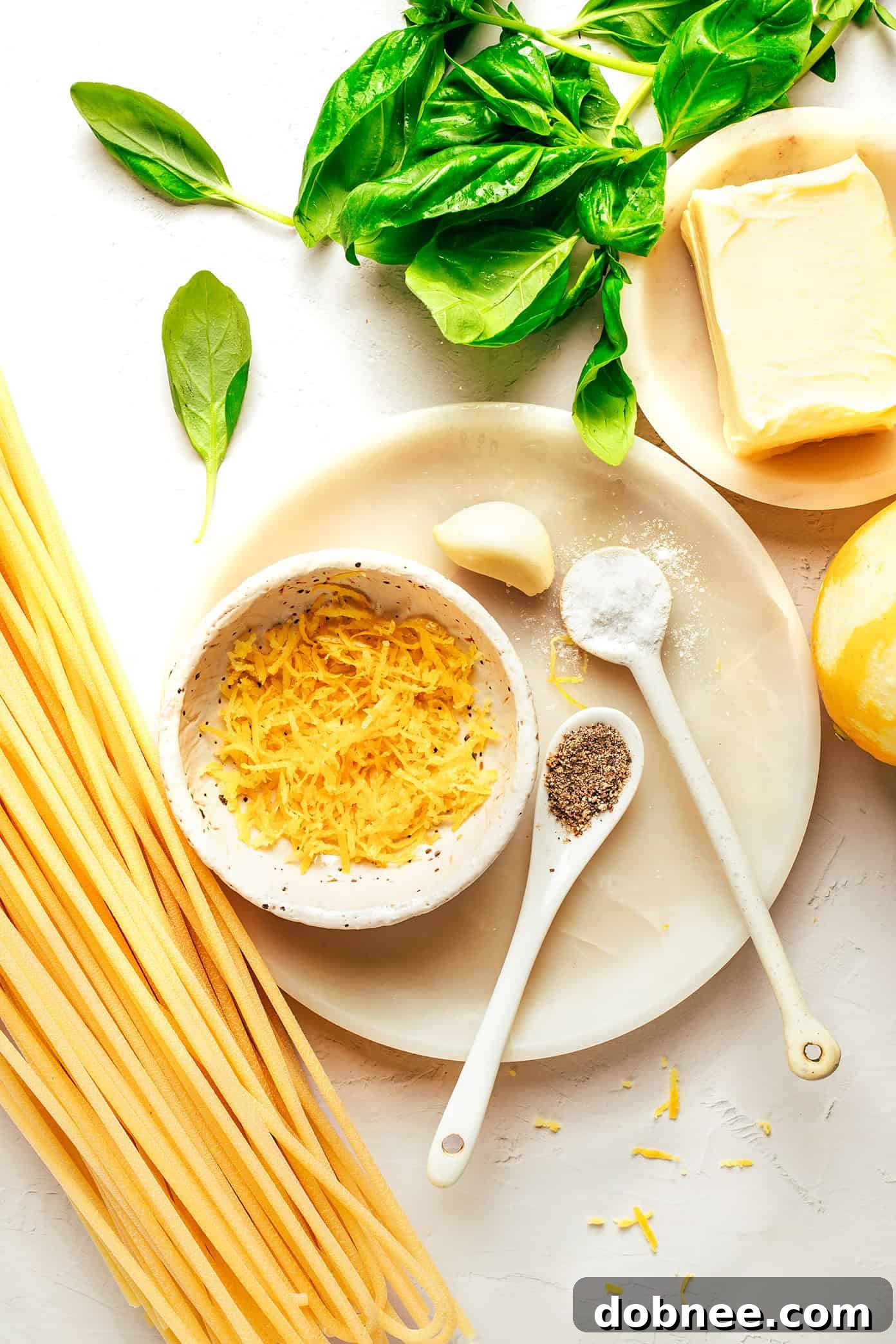 A selection of fresh ingredients laid out on a wooden board, including fresh basil leaves, a block of butter, a lemon, garlic cloves, and a variety of pasta shapes, ready for making lemon basil butter pasta.