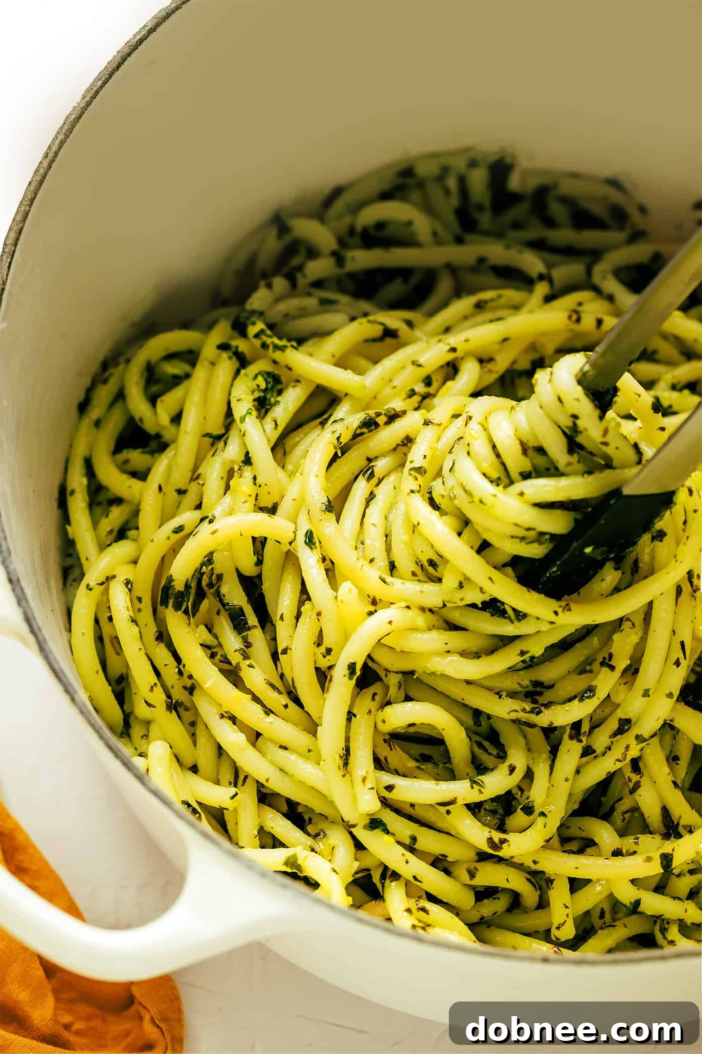 Lemon basil butter pasta being tossed with tongs in a large pot, the vibrant green sauce coating the bucatini strands evenly.