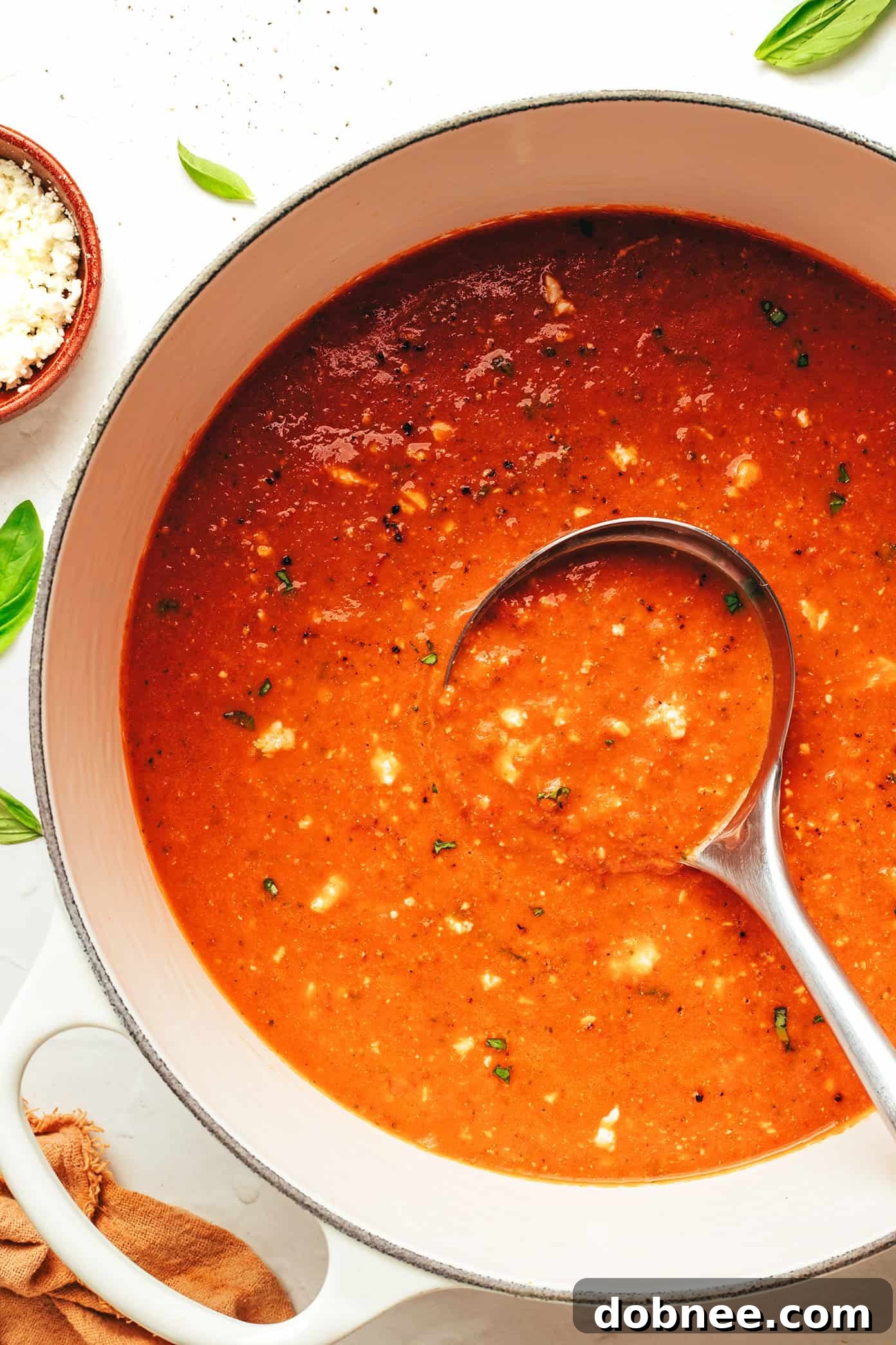 A bowl of creamy tomato feta soup garnished with fresh basil and more crumbled feta, with a spoon dipping in.