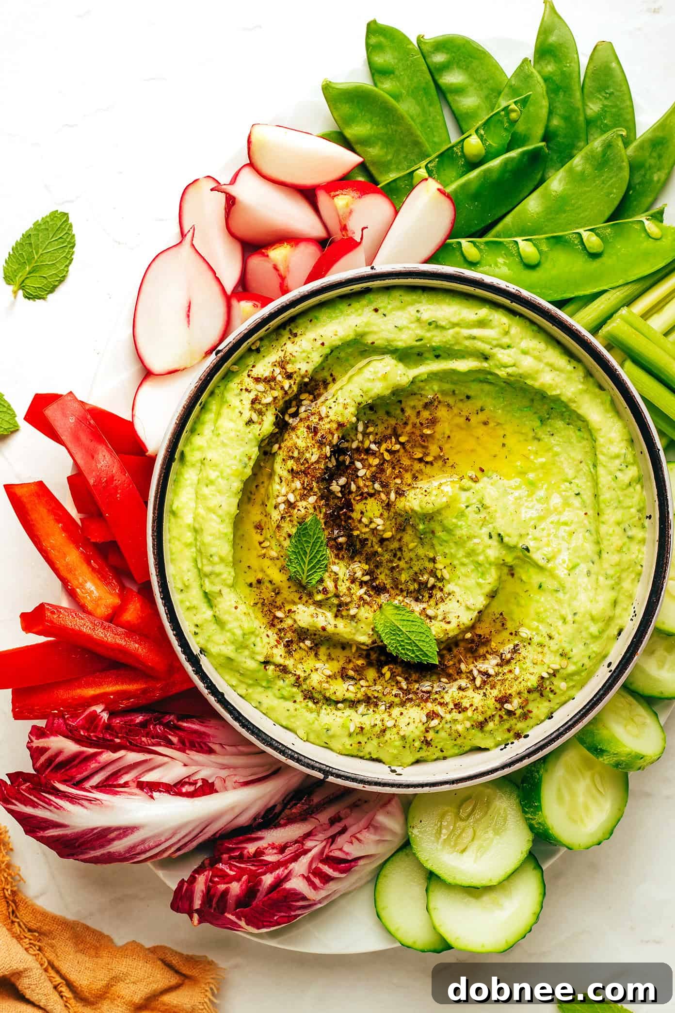 Vibrant Mint & Pea Hummus 2 Bright green mint pea hummus in a bowl, garnished with fresh mint leaves and a drizzle of olive oil, surrounded by pita bread and veggie sticks.
