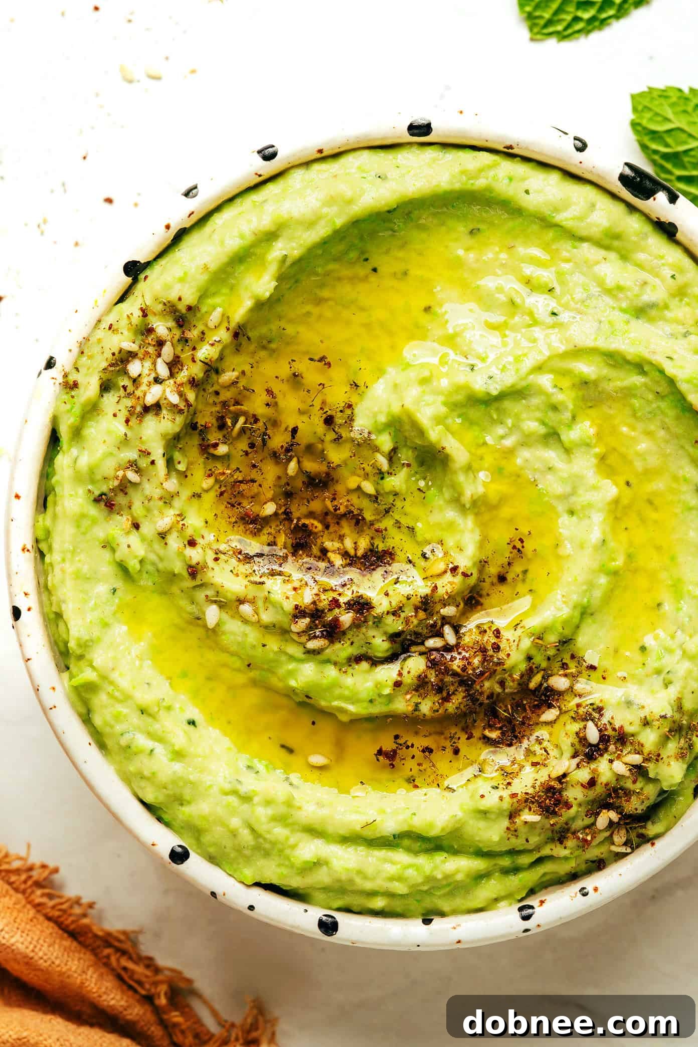 Vibrant Mint & Pea Hummus 7 A close-up of mint pea hummus being swirled with a spoon, showing its creamy texture, with a garnish of a single fresh mint leaf and a slight drizzle of olive oil.