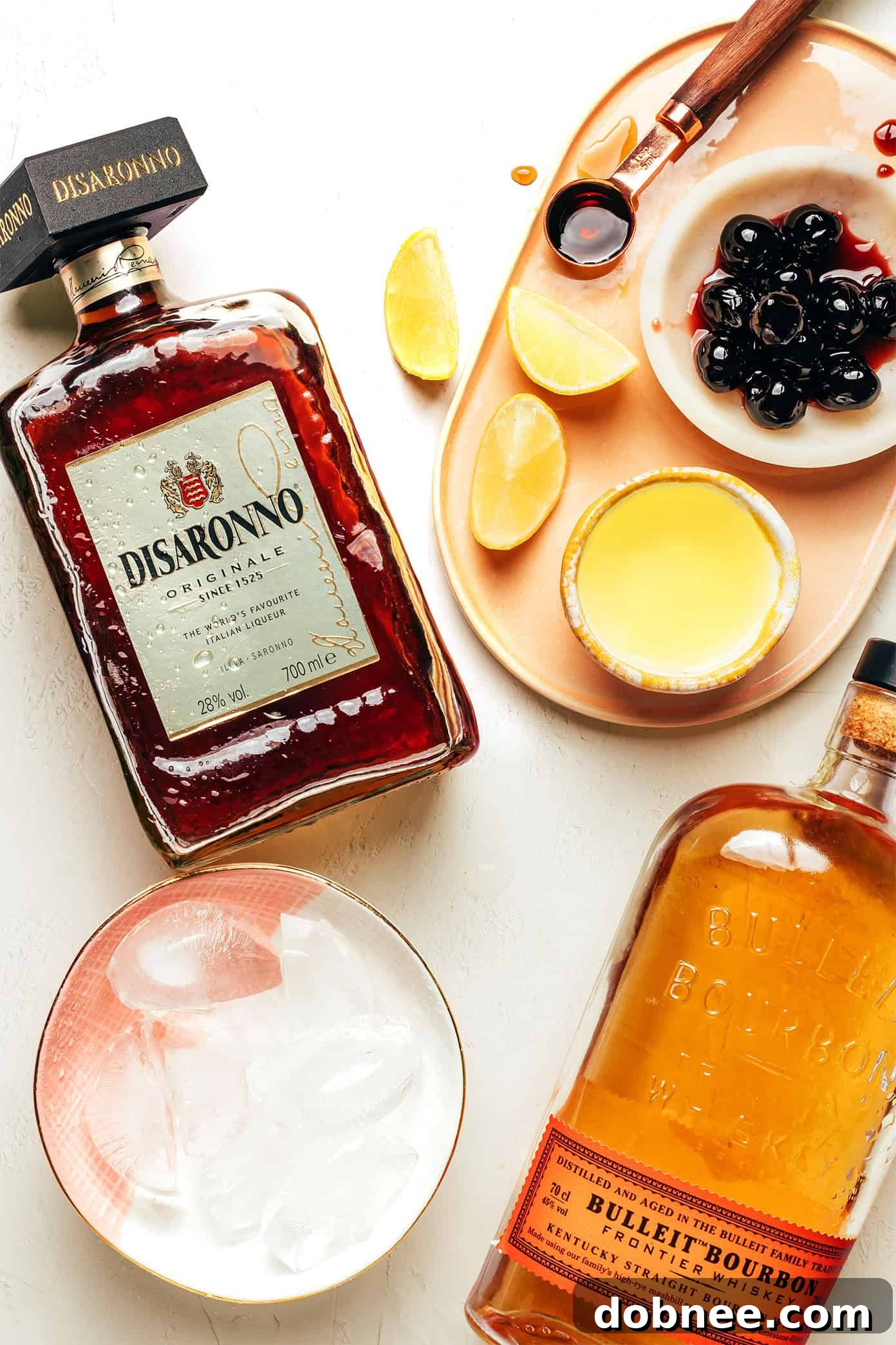 This classic Amaretto Sour recipe is easy to make with bourbon, lemon juice, Amaretto and egg white. Feel free to add additional sweetener or bitters, if you'd like, and garnish with a lemon twist and maraschino cherry. It's always such a cozy and sophisticated cocktail! | gimmesomeoven.com Amaretto Sour ingredients laid out: Amaretto, bourbon, lemon, maple syrup, egg white, and bitters.