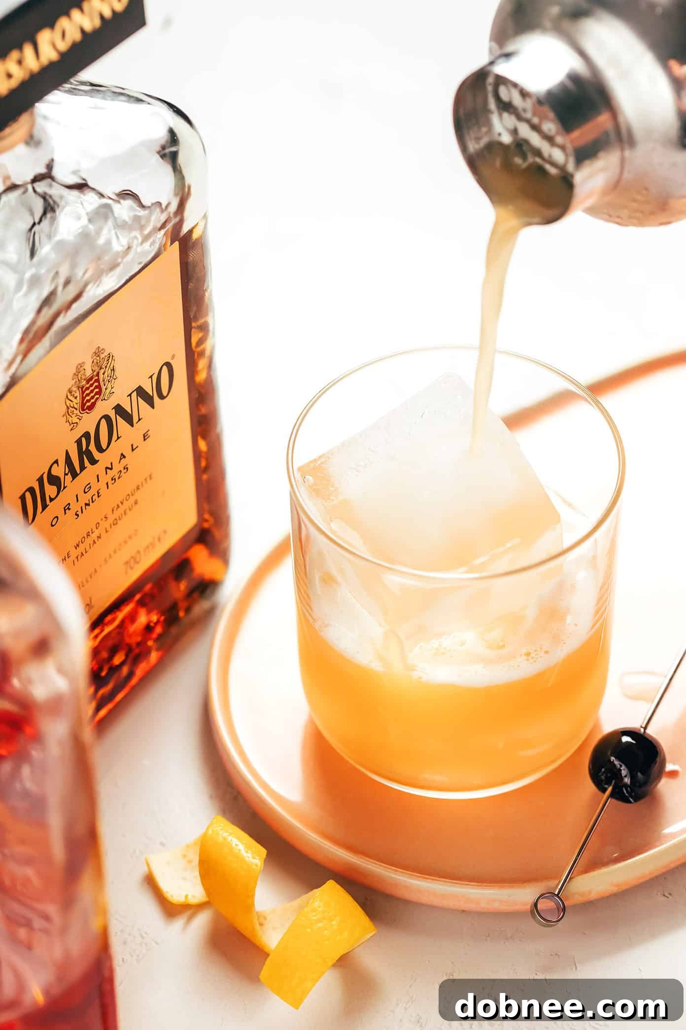 This classic Amaretto Sour recipe is easy to make with bourbon, lemon juice, Amaretto and egg white. Feel free to add additional sweetener or bitters, if you'd like, and garnish with a lemon twist and maraschino cherry. It's always such a cozy and sophisticated cocktail! | gimmesomeoven.com Hands preparing an Amaretto Sour cocktail by shaking ingredients in a cocktail shaker.