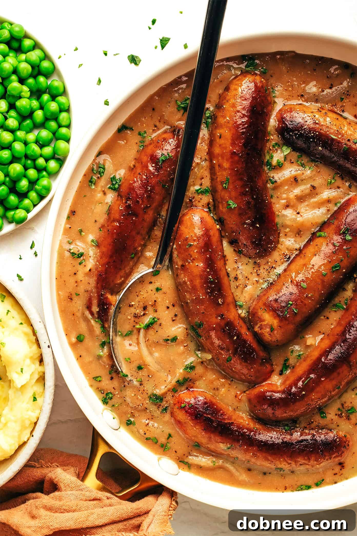 British Comfort Classic 5 Sausages and gravy being spooned onto a bed of mashed potatoes, with fresh parsley garnish.