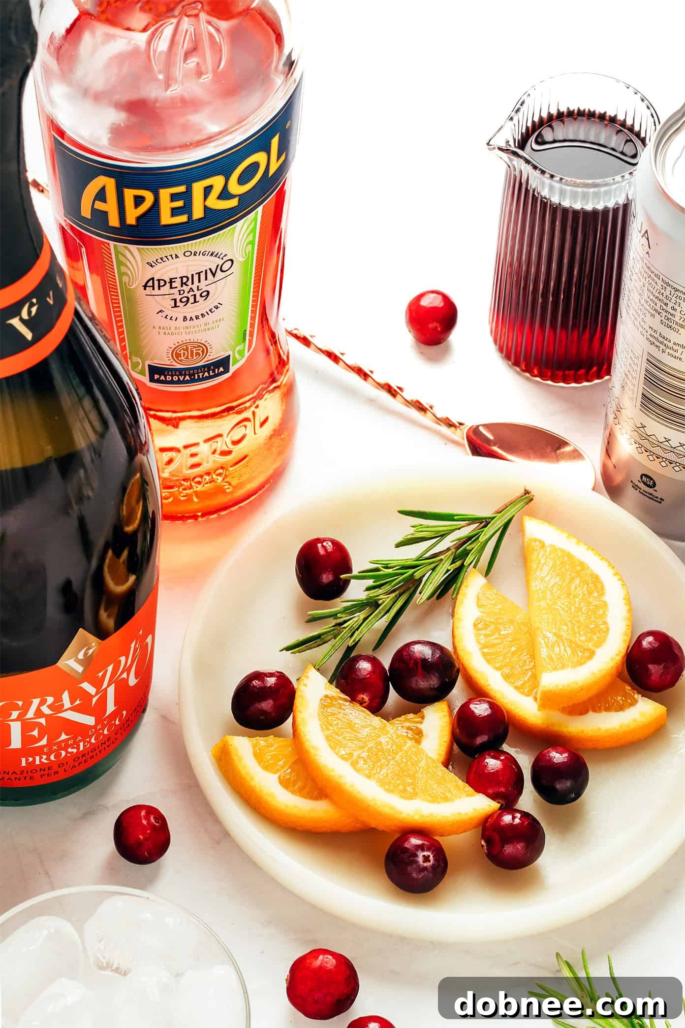 Ingredients for Cranberry Aperol Spritz laid out: Aperol bottle, Prosecco bottle, cranberry juice, soda water, fresh cranberries, orange slices, and rosemary sprigs.